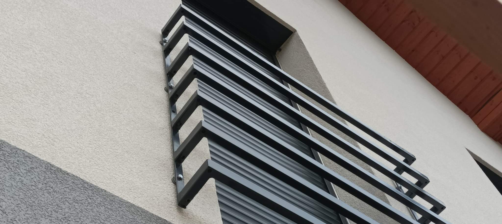 Nowoczesna, grafitowa balustrada balkonowa z poziomych profili, zamontowana na elewacji budynku z widocznymi śrubami mocującymi. Ujęcie z dołu, podkreślające geometrię.