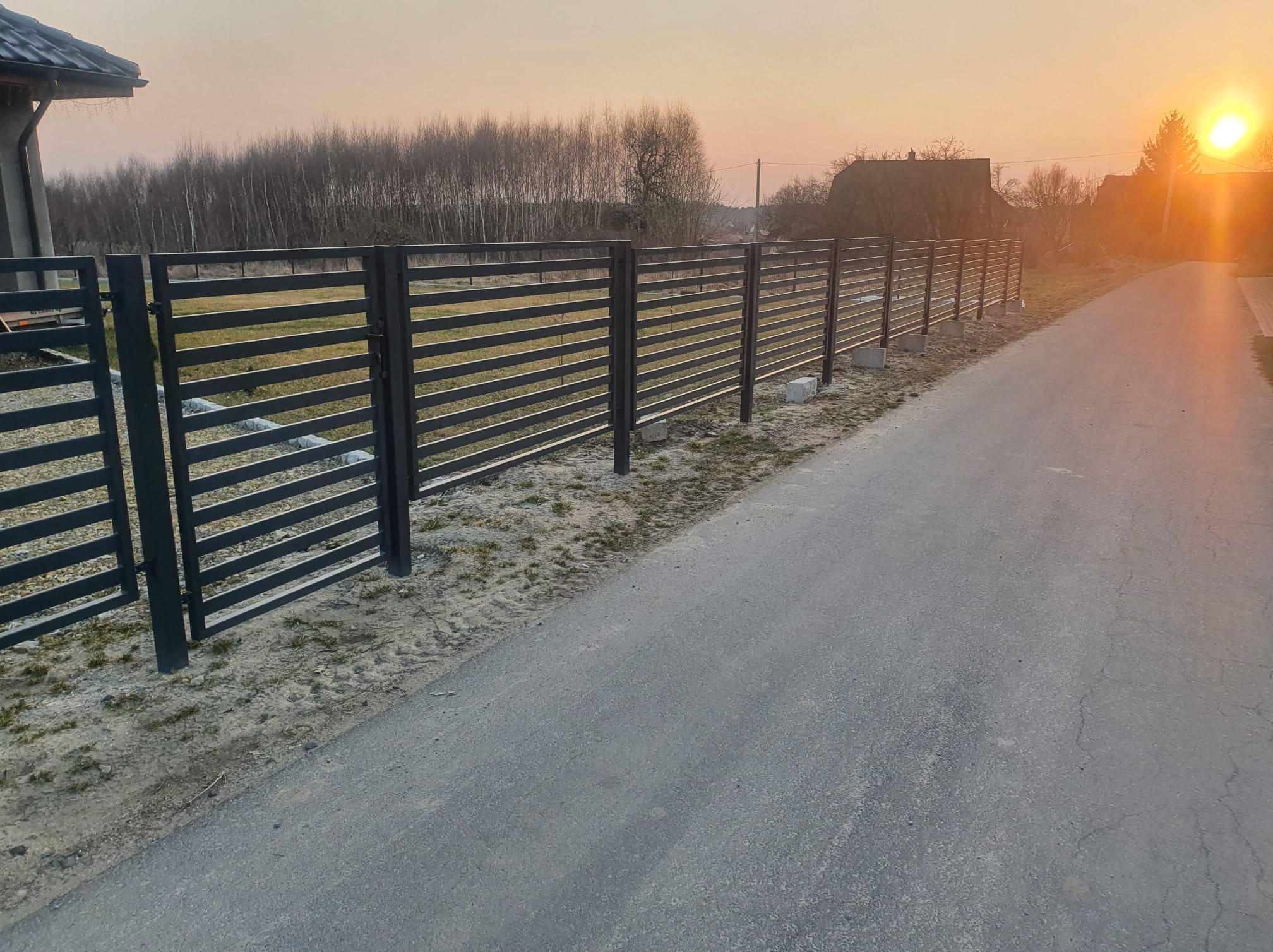 Nowoczesne ogrodzenie metalowe z poziomymi panelami, widok z perspektywy drogi, w tle drzewa i zachodzące słońce. Minimalistyczny design, szara kolorystyka.