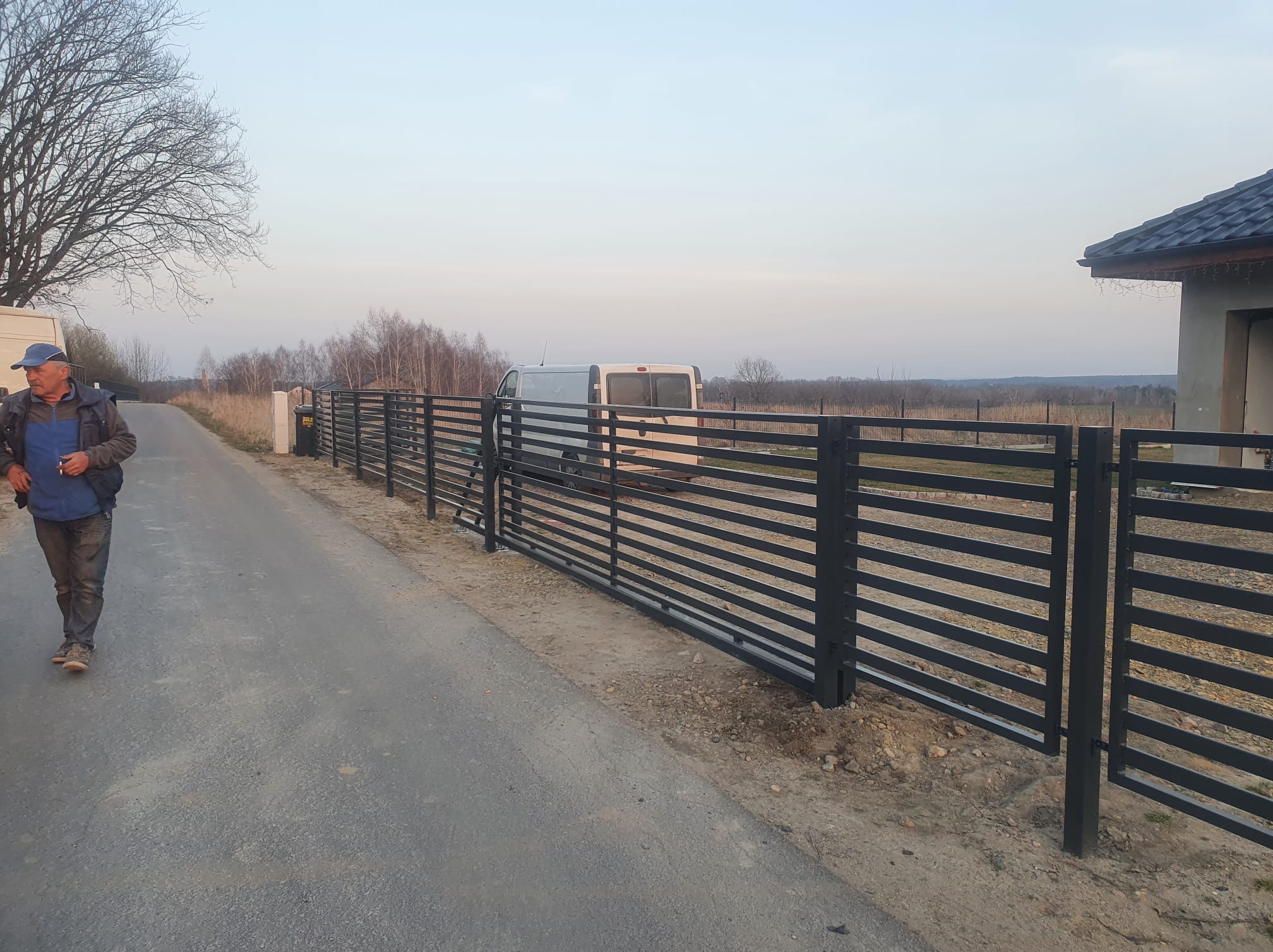 Nowoczesne, poziome ogrodzenie metalowe w kolorze antracytowym, oddzielające posesję od drogi. Widoczny fragment domu i osoba idąca chodnikiem.