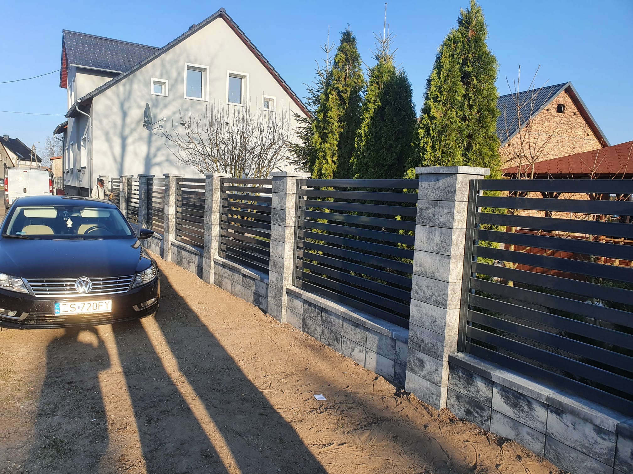 Nowoczesne ogrodzenie poziome z szarymi, betonowymi słupkami i czarnymi, metalowymi przęsłami, na tle domu i zielonych drzew. Ciemny samochód osobowy zaparkowany obok.