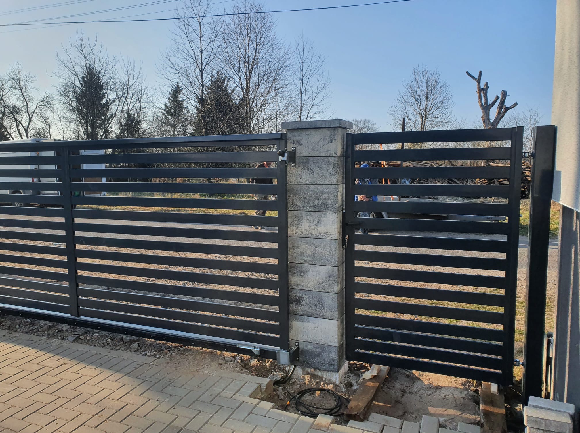 Nowoczesna, przesuwna brama wjazdowa z poziomych, czarnych profili, osadzona na betonowych słupkach. Widoczne elementy automatyki i kostka brukowa.