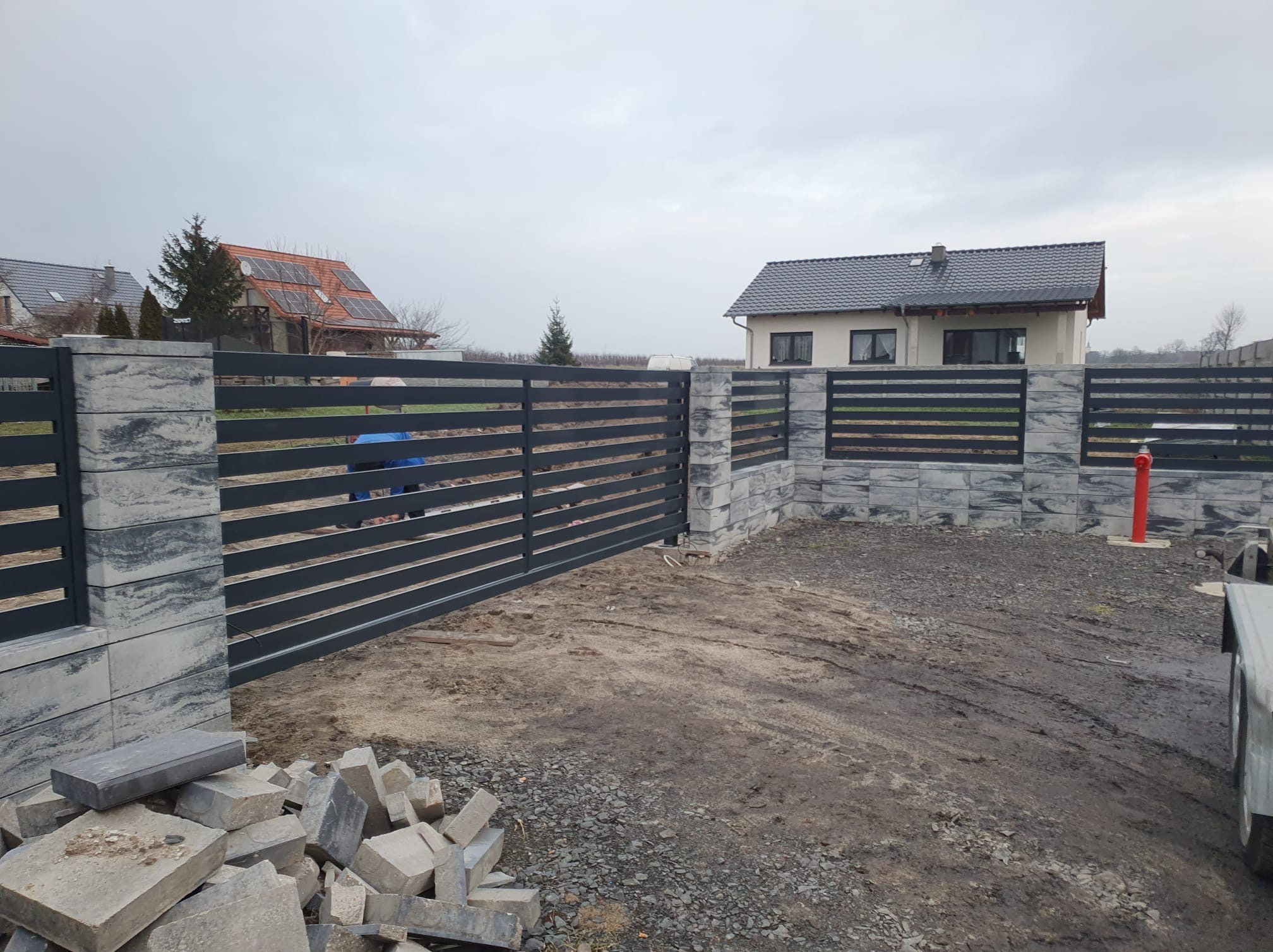 Nowoczesna, pozioma brama wjazdowa z szarego metalu, osadzona na kamiennych słupkach. W tle domy jednorodzinne i pochmurne niebo. Część kamieni leży na ziemi.