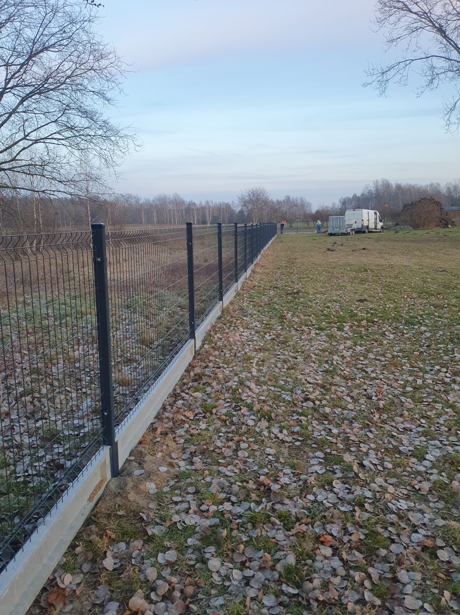 Długi, panelowy płot z podmurówką, ciągnący się wzdłuż działki pokrytej liśćmi. W tle widoczny las i ekipa montażowa z samochodem dostawczym.