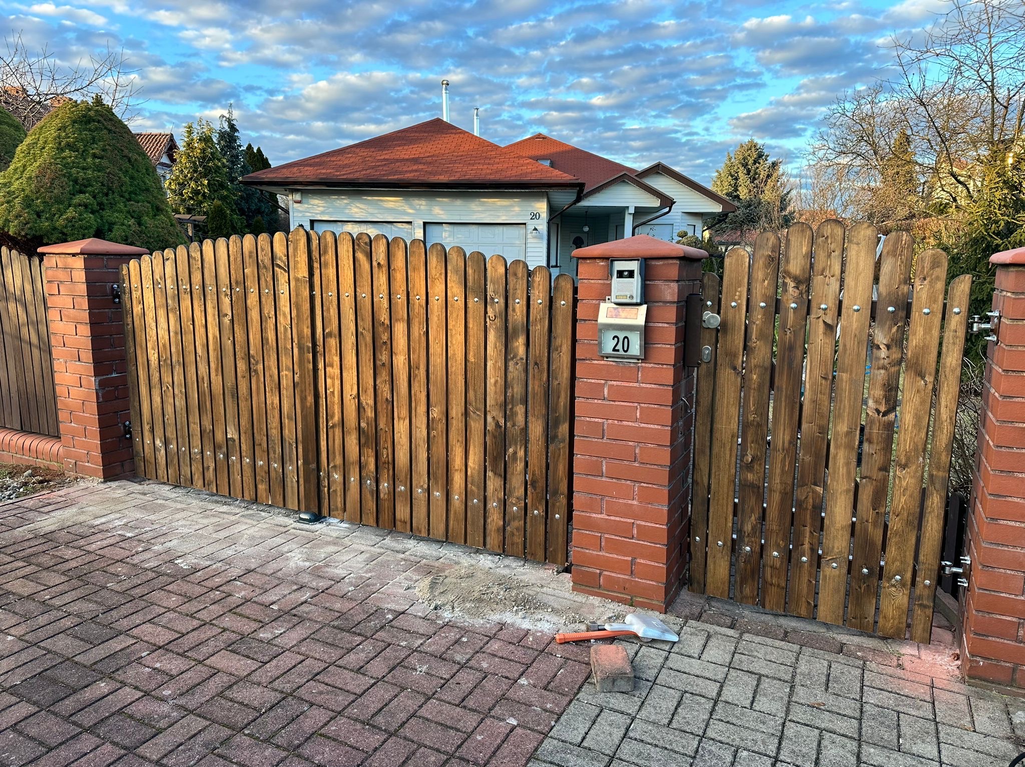 Drewniana brama wjazdowa z zaokrąglonymi sztachetami, osadzona między ceglanymi słupkami z numerem domu. Widoczny fragment posesji i kostka brukowa.