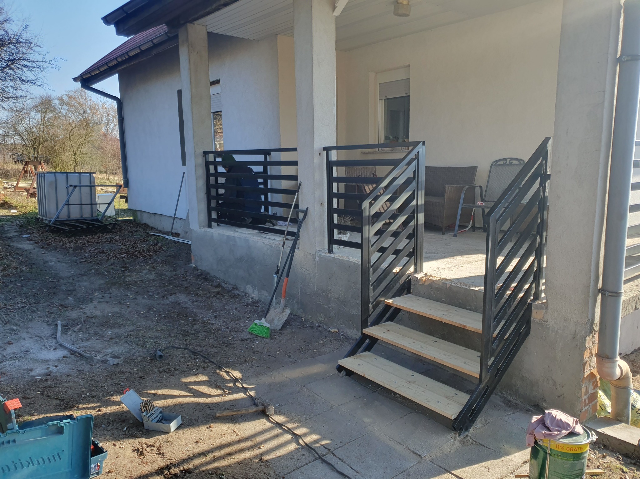 Nowoczesna, czarna balustrada schodowa z drewnianymi stopniami, zamontowana przy wejściu do domu. Widoczne narzędzia i akcesoria montażowe. Słoneczny dzień.