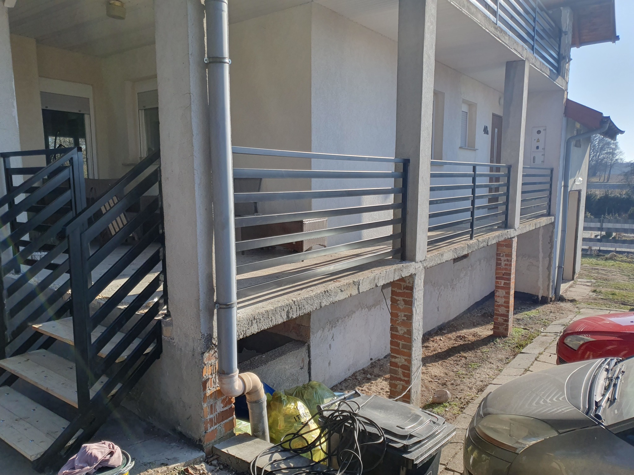 Nowoczesna balustrada balkonowa z poziomych profili, zamontowana przy budynku z widocznymi schodami wejściowymi i elementami wykończeniowymi. Jasne tło, słoneczny dzień.