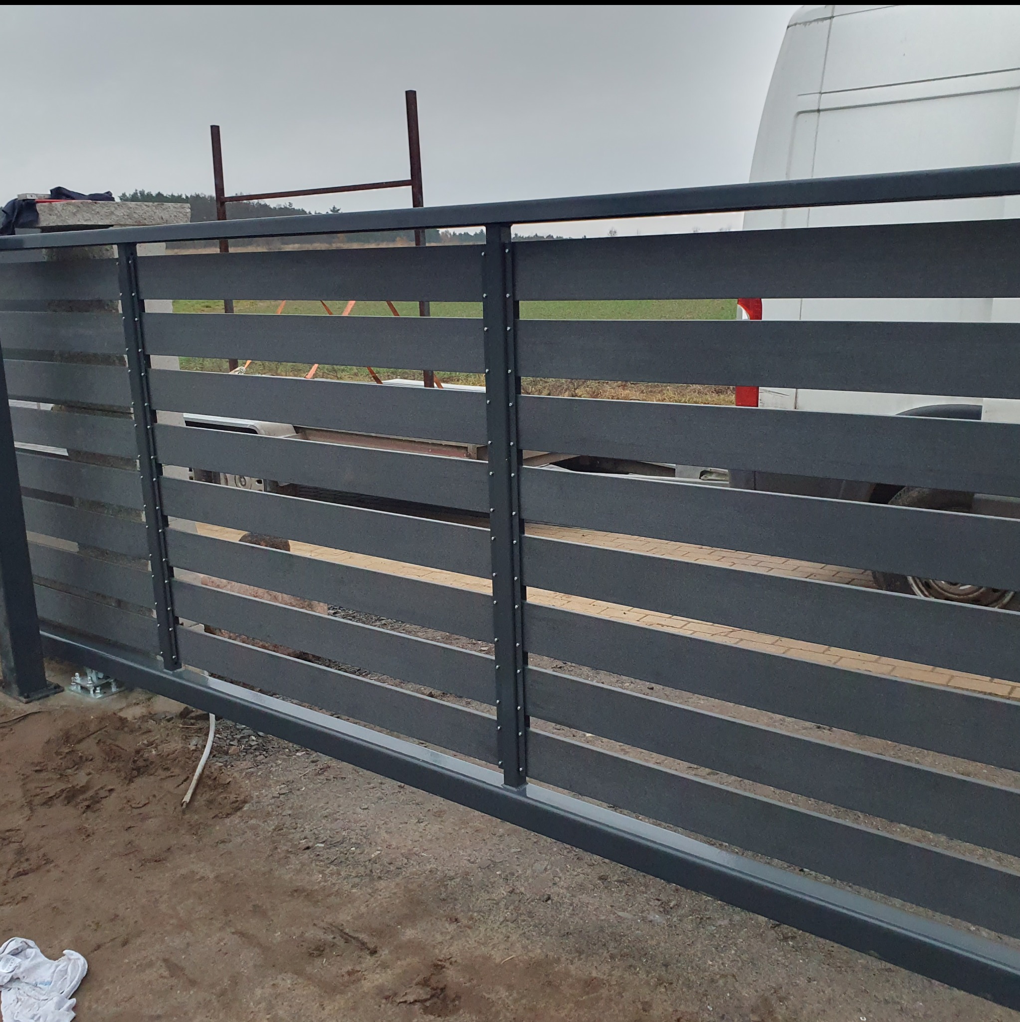 Nowoczesna, przesuwna brama wjazdowa z poziomych, szarych paneli, zamontowana na posesji. Widoczne elementy konstrukcyjne i podłoże.