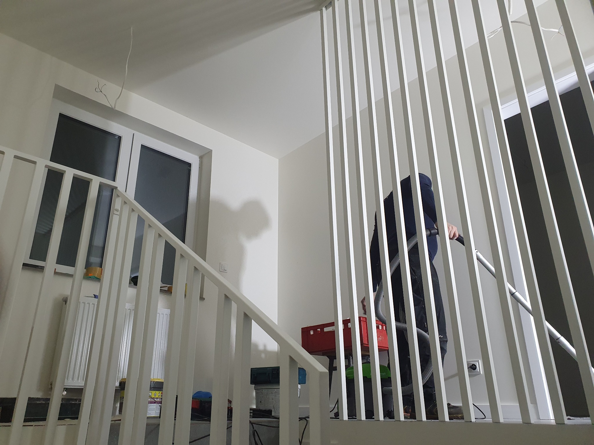 Białe, pionowe balustrady schodowe w jasnym wnętrzu. Osoba odkurza podłogę, widoczna przez szpary balustrad. W tle okno i grzejnik.