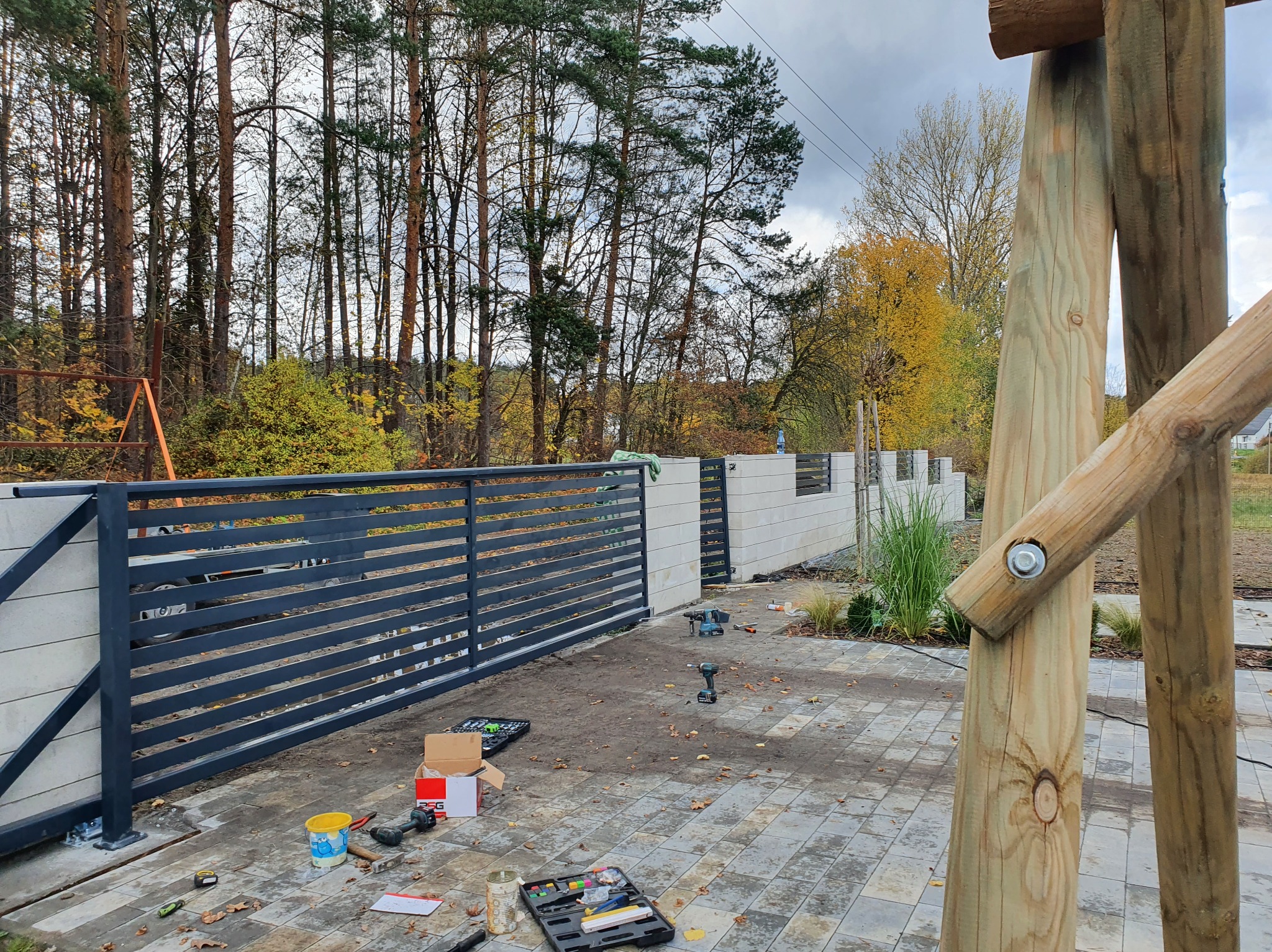 Montaż nowoczesnej, poziomej bramy wjazdowej z szarego metalu. Widoczne narzędzia i elementy konstrukcyjne w trakcie instalacji na tle jesiennego krajobrazu.