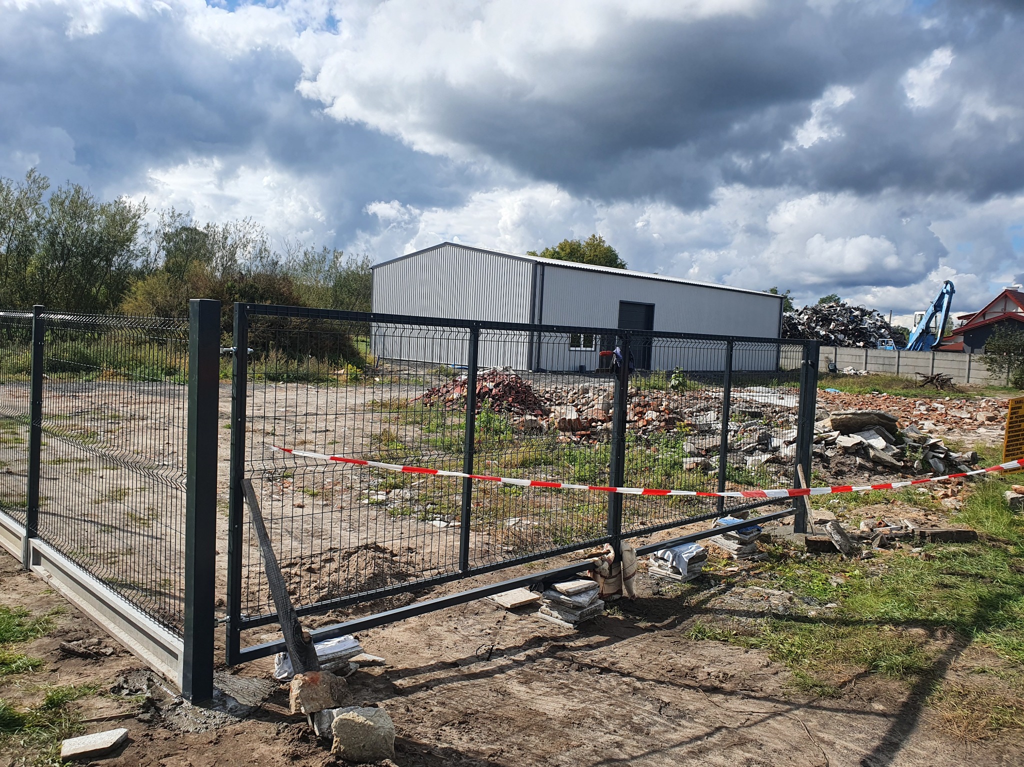 Czarna metalowa brama wjazdowa z siatki, w trakcie montażu, z widocznymi elementami betonowymi u podstawy. W tle budynek przemysłowy i pochmurne niebo.
