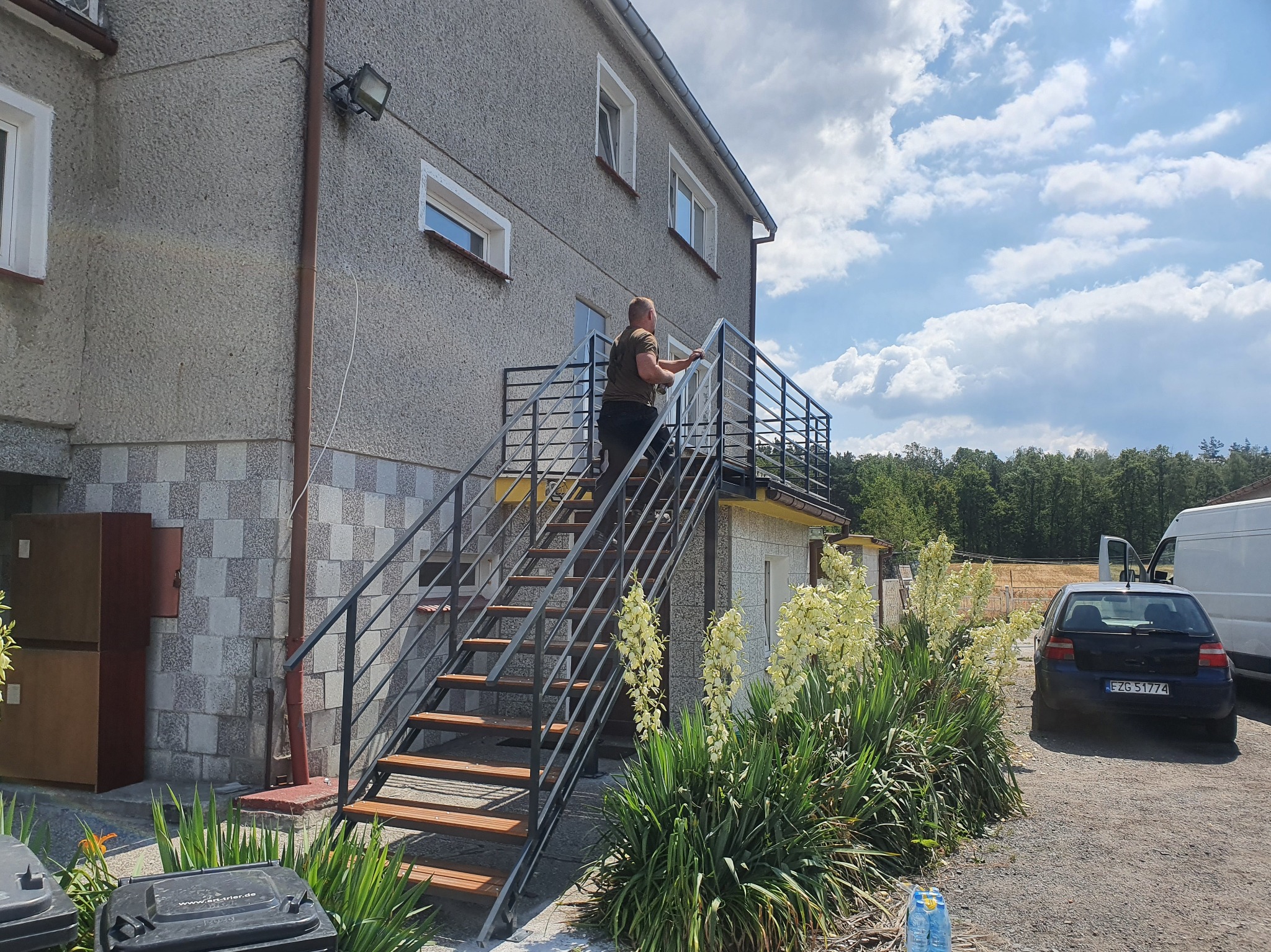 Metalowe schody z drewnianymi stopniami prowadzą na balkon budynku. Mężczyzna czyści balustradę. W tle zaparkowane auto i bus. Na pierwszym planie kwitnące juki.