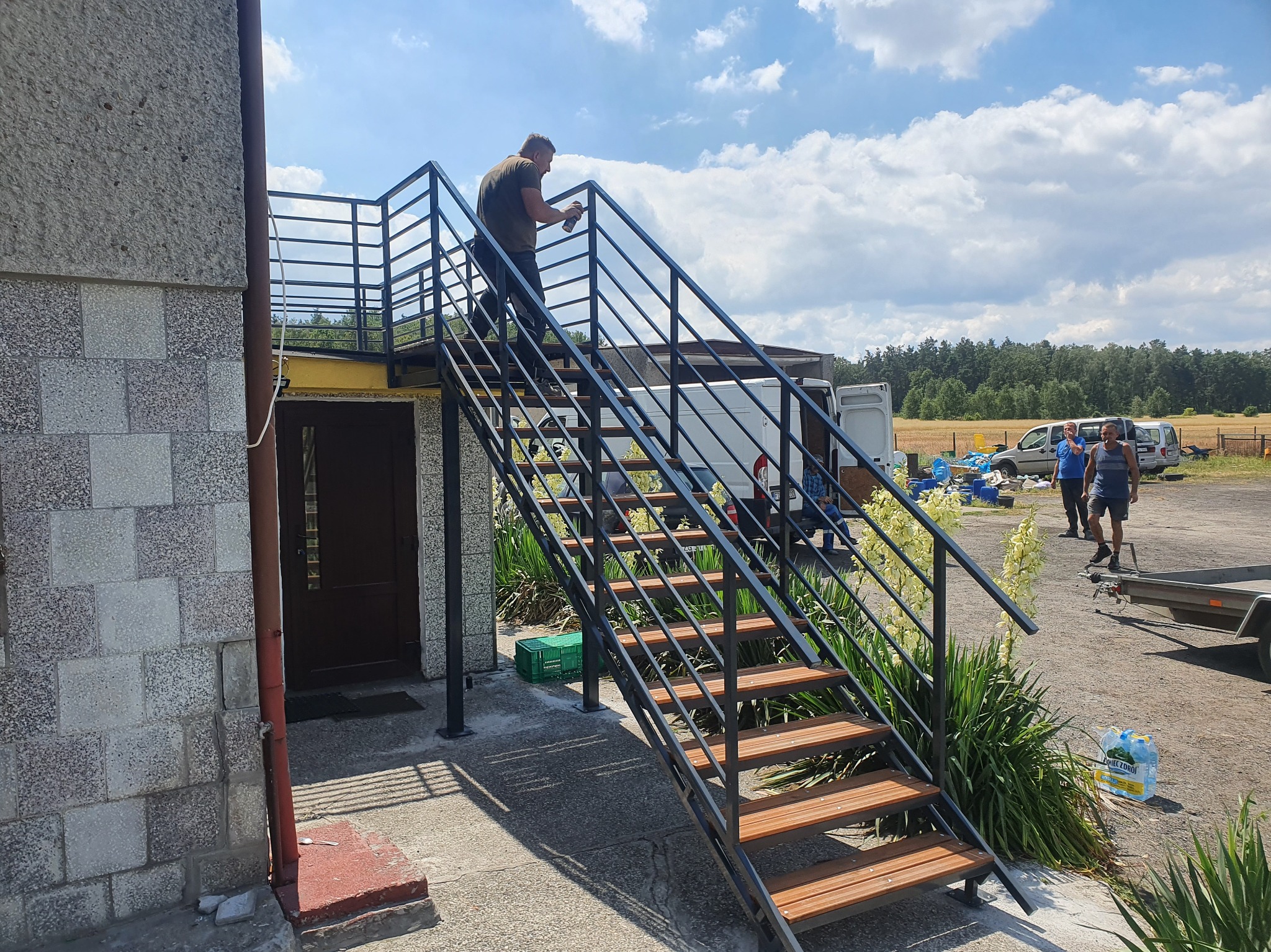 Zewnętrzne schody metalowe z drewnianymi stopniami, malowane przez mężczyznę. W tle budynek, pojazdy i osoby. Dzień słoneczny, błękitne niebo z chmurami.