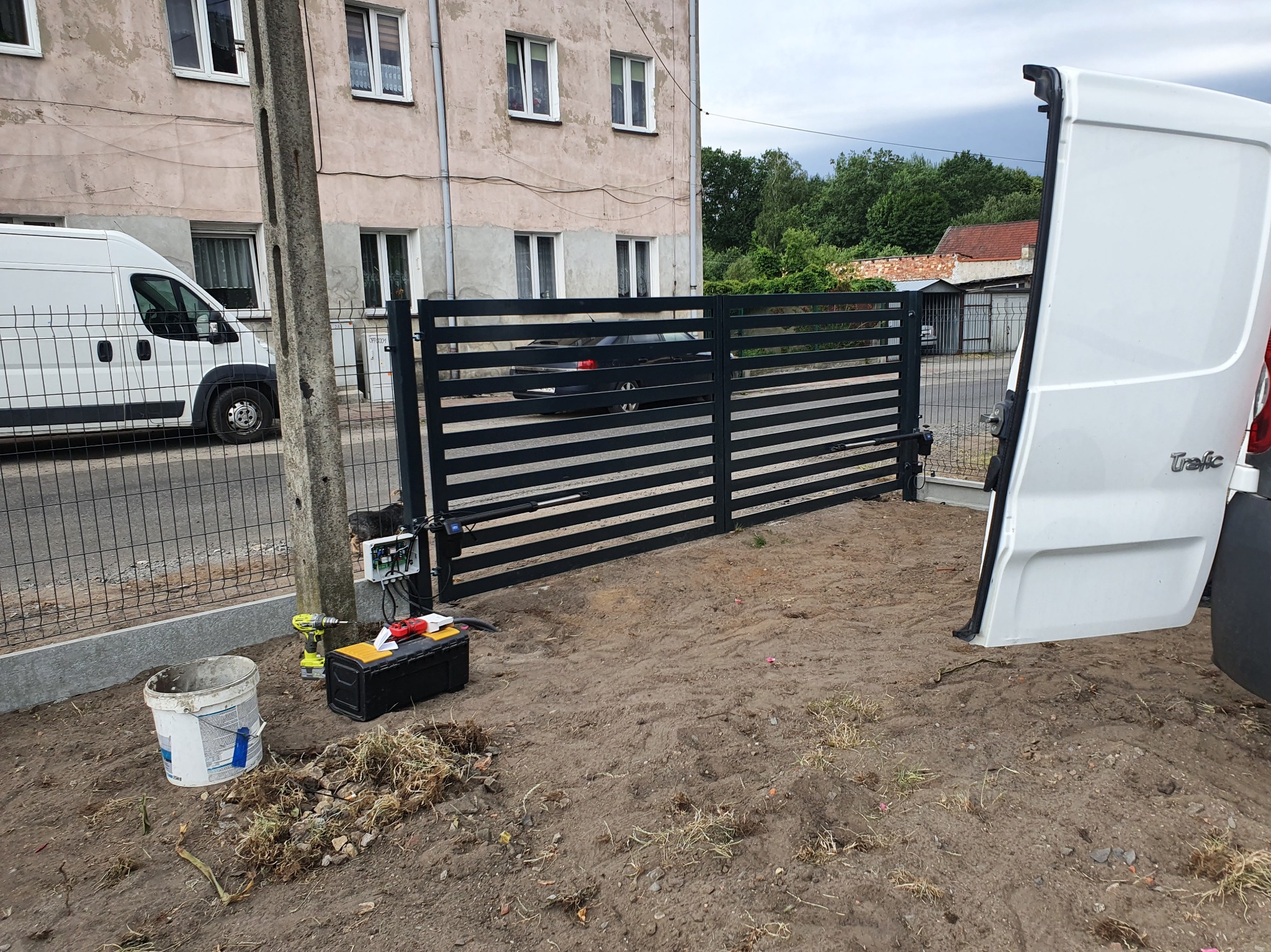 Czarna, pozioma brama wjazdowa z automatyką, częściowo otwarta, z narzędziami montażowymi obok. W tle budynek i biały samochód dostawczy.
