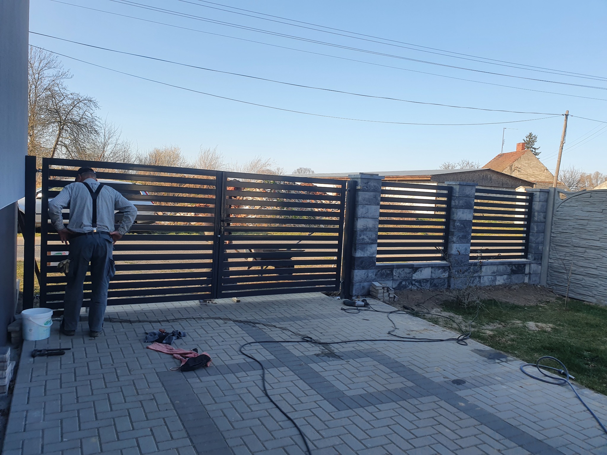 Nowoczesna brama wjazdowa z poziomych profili, częściowo zamontowana. Widoczny monter w szelkach i narzędzia na kostce brukowej. Słupki z szarego kamienia.