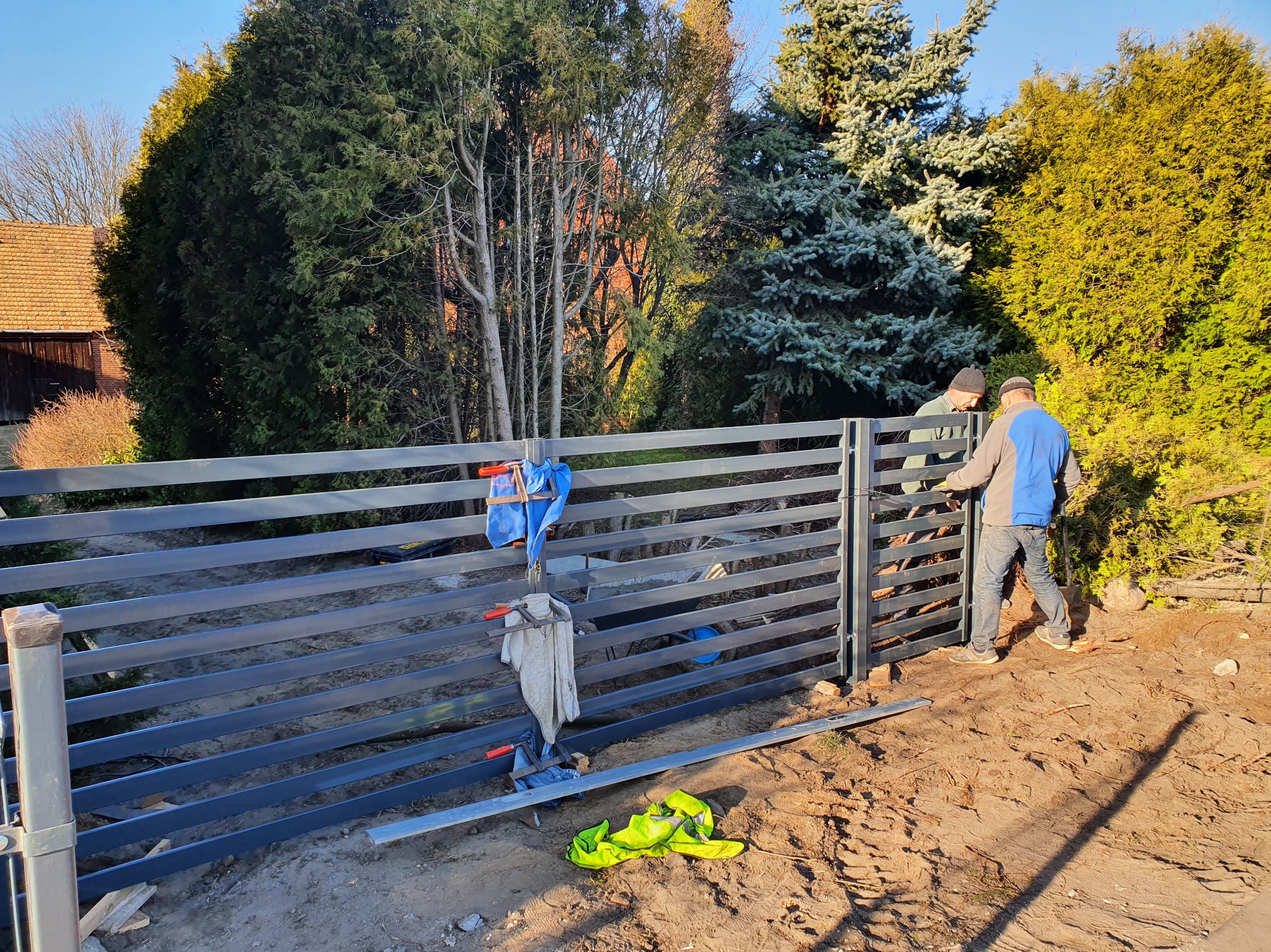 Montaż przęsła ogrodzeniowego z poziomych profili stalowych przez dwóch monterów. Widoczne narzędzia i odzież robocza. W tle zieleń drzew i budynek.