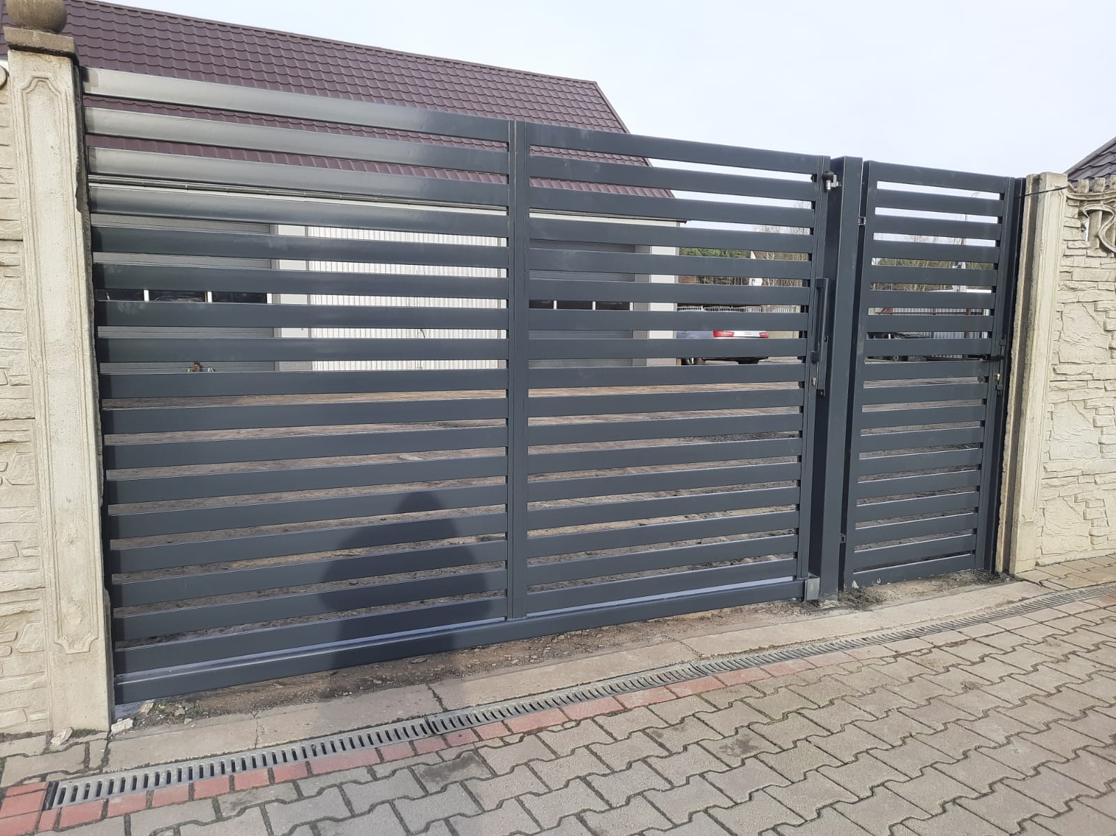 Nowoczesna, przesuwna brama wjazdowa z poziomych, szarych profili, zamontowana między dwoma kamiennymi słupkami. Widoczny fragment posesji i kostka brukowa.