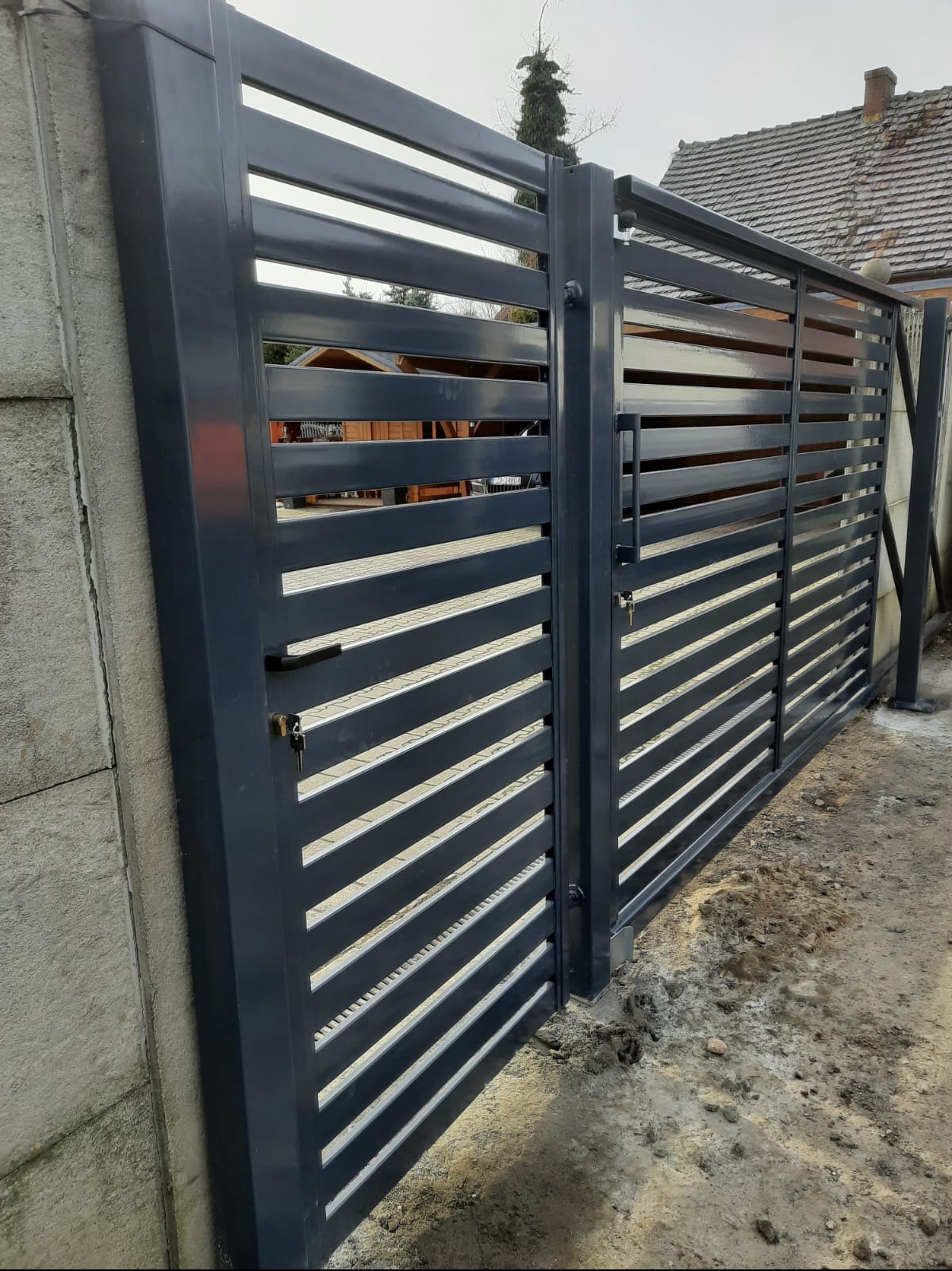 Nowoczesna, pozioma brama wjazdowa z metalowymi lamelami w kolorze grafitowym, zamontowana przy betonowym słupku. Widoczny zamek i uchwyt.