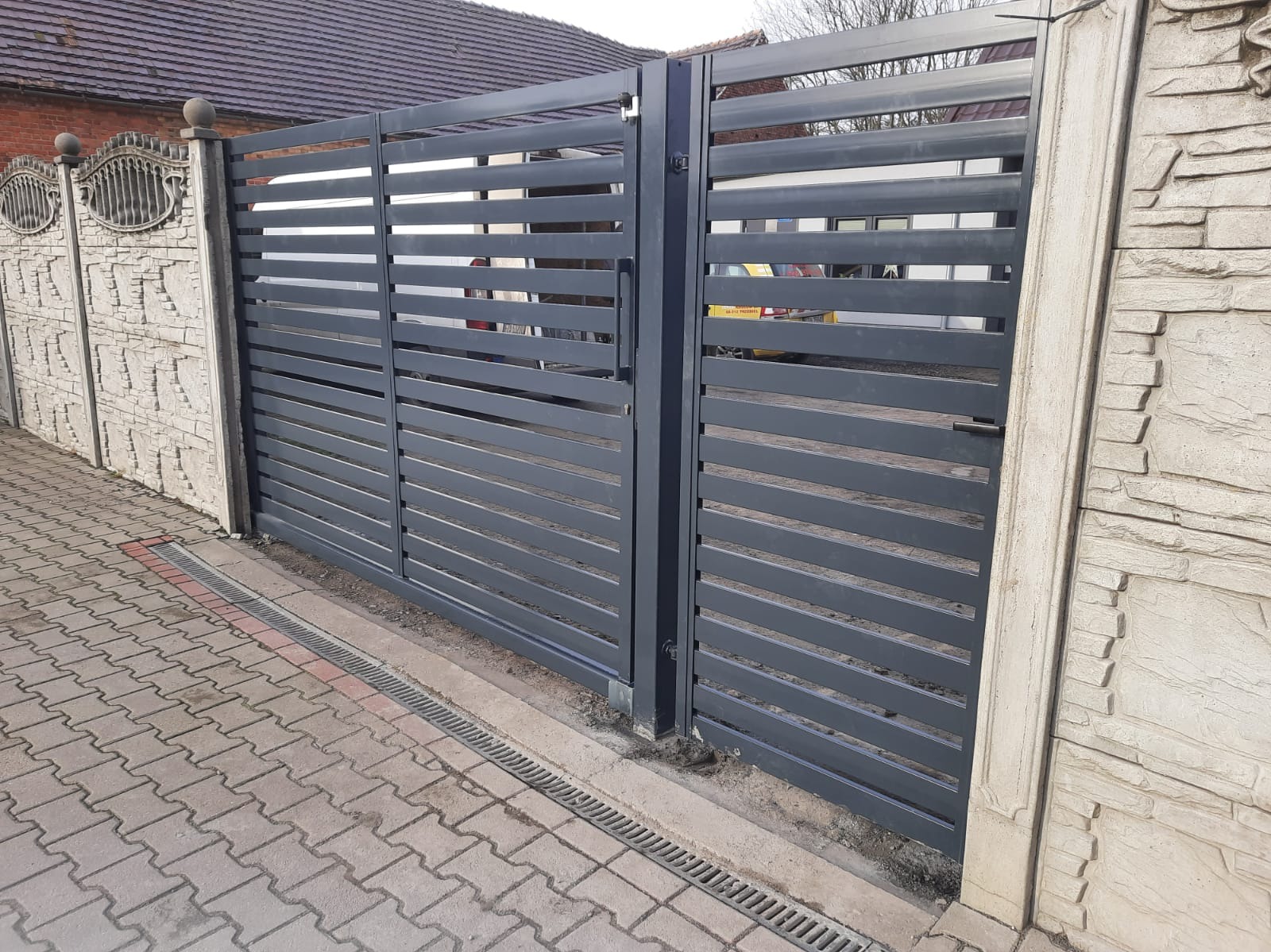 Nowoczesna, przesuwna brama wjazdowa z poziomych profili, szara, zamontowana między ozdobnymi, betonowymi słupkami. Widoczna kostka brukowa i odpływ liniowy.