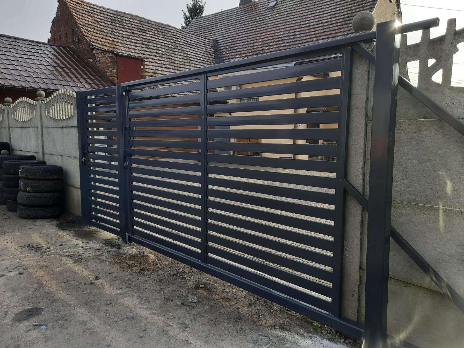 Nowoczesna, pozioma brama przesuwna w kolorze grafitowym, z widocznymi elementami konstrukcyjnymi i betonowym ogrodzeniem w tle. Widoczne opony przy ogrodzeniu.
