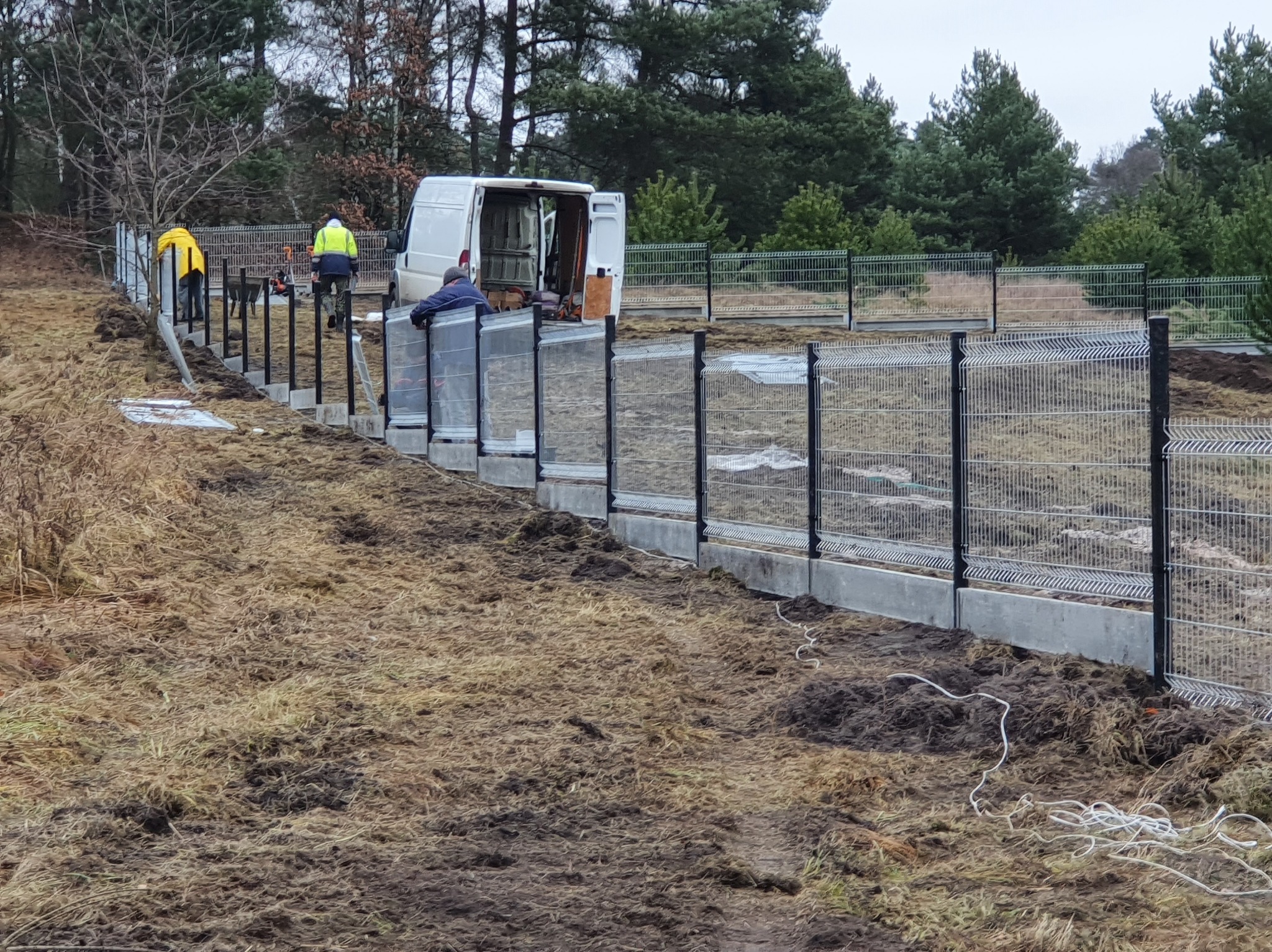 Montaż ogrodzenia panelowego na betonowej podmurówce. Widoczni pracownicy w odblaskowych ubraniach i biały bus w tle. Teren trawiasty, częściowo rozkopany.