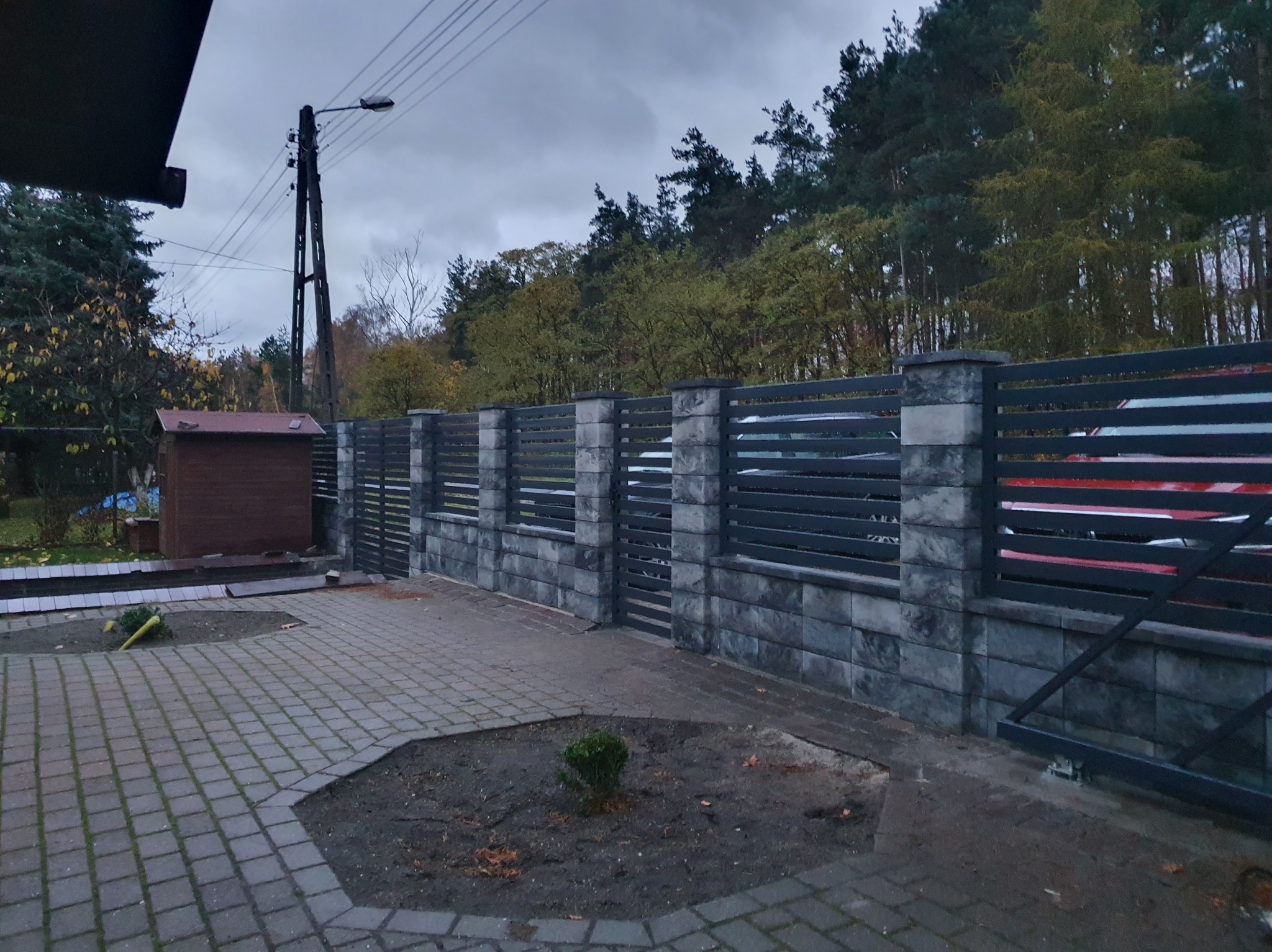 Nowoczesne ogrodzenie z szarego kamienia i metalowych paneli, widok z zewnątrz posesji, brama wjazdowa częściowo otwarta, jesienne otoczenie.