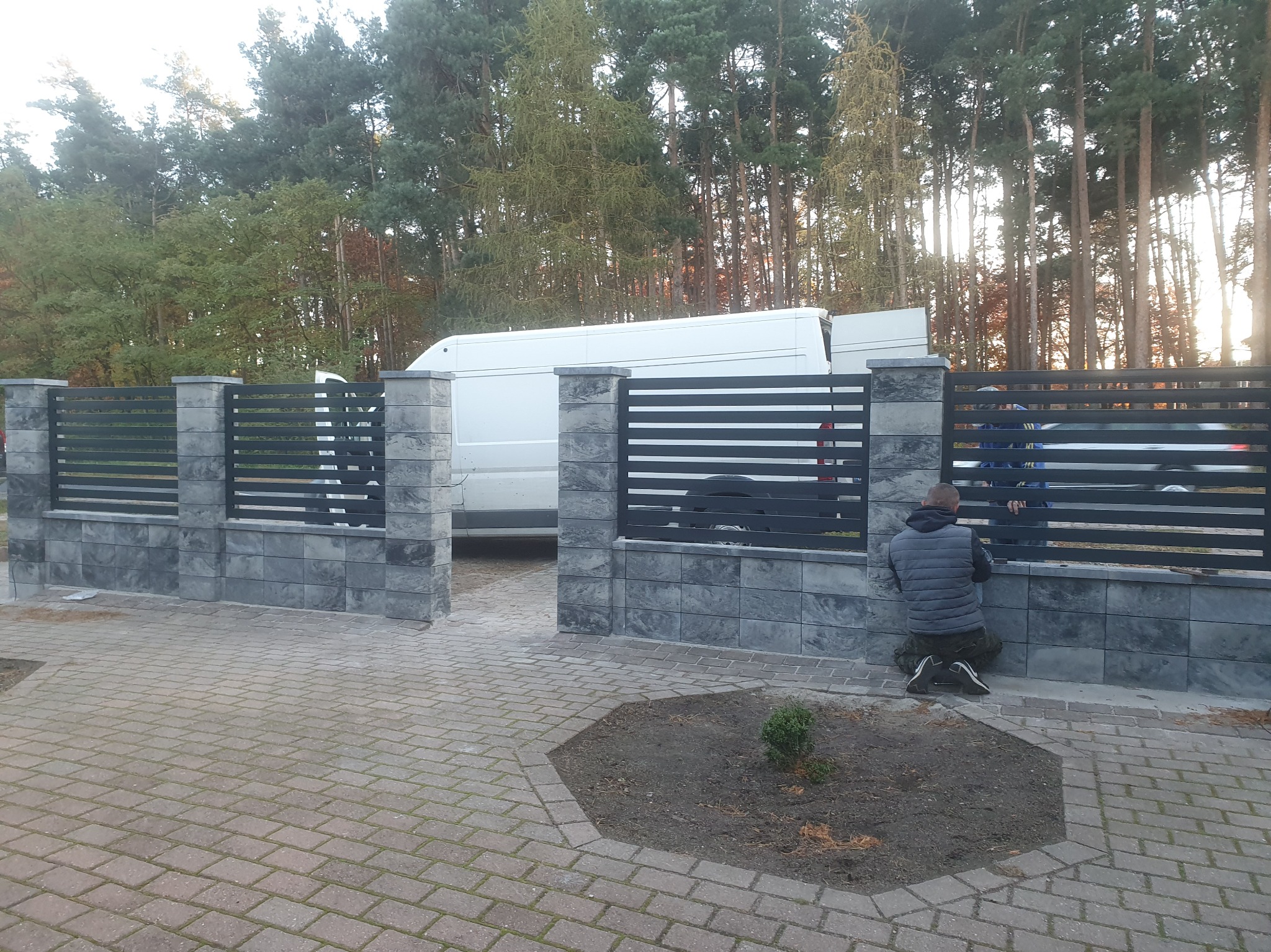 Nowoczesne ogrodzenie z szarych bloczków i poziomych, czarnych przęseł, z białym busem w tle i osobą montującą elementy. Widok z zewnątrz posesji.