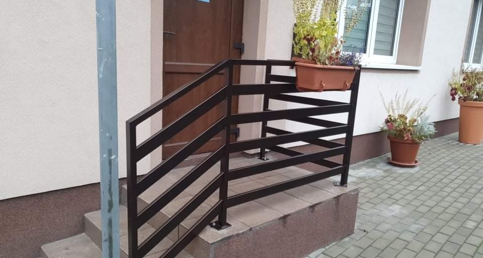 Nowoczesna, brązowa balustrada schodowa z poziomymi szczeblami, z donicą z kwiatami, przy wejściu do budynku z brązowymi drzwiami i oknem.