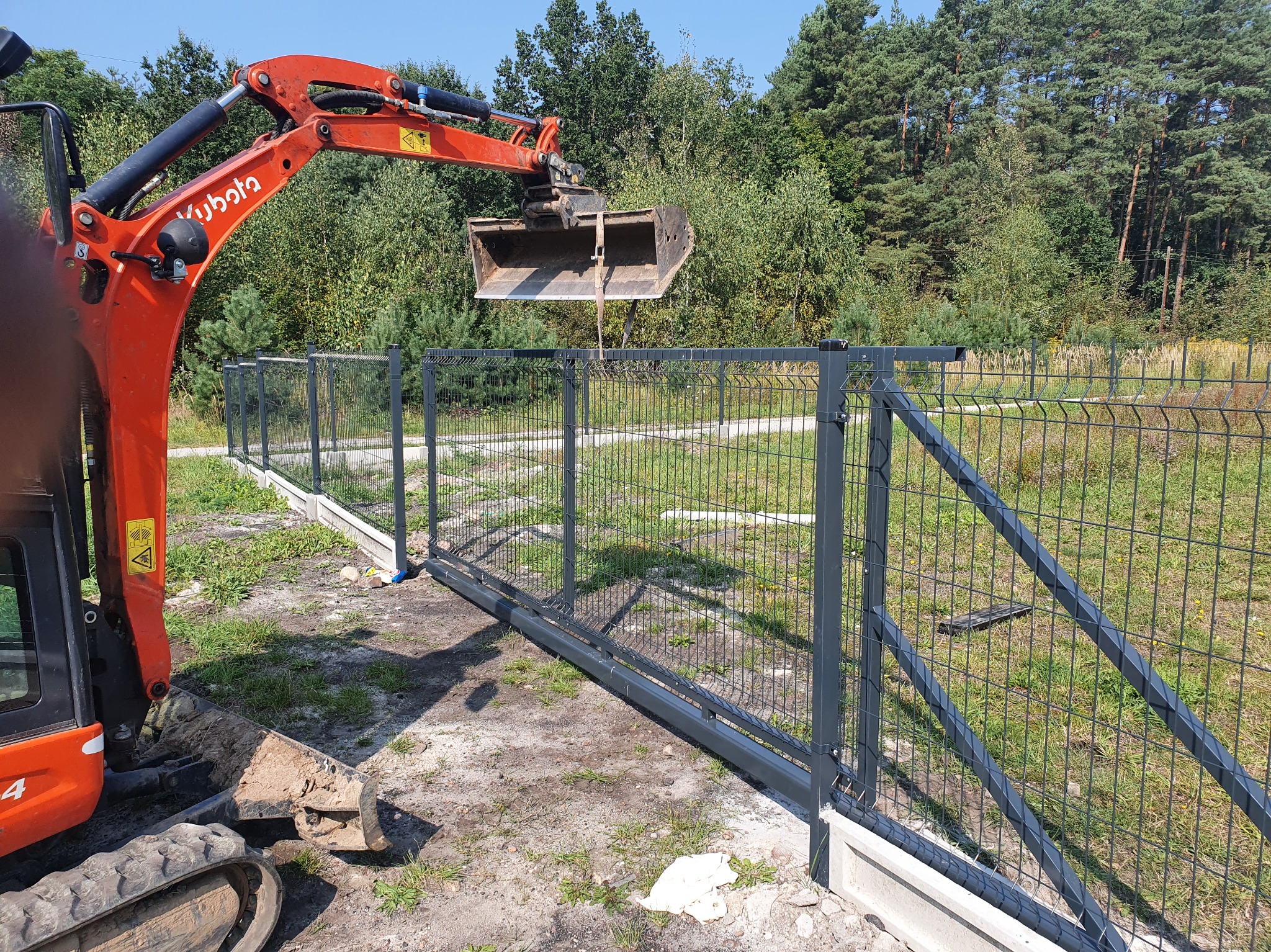 Montaż ogrodzenia panelowego z bramą przesuwną, wsparty przez pomarańczową koparkę Kubota. Widoczne betonowe fundamenty i leśne tło.