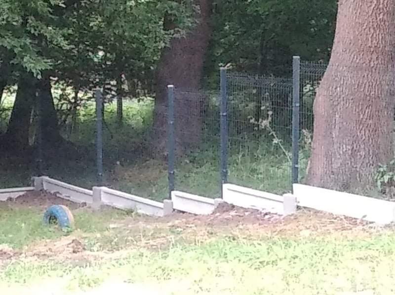 Fragment montażu ogrodzenia panelowego z podmurówką betonową na nierównym terenie, widoczne drzewa w tle. Etap budowy ogrodzenia.