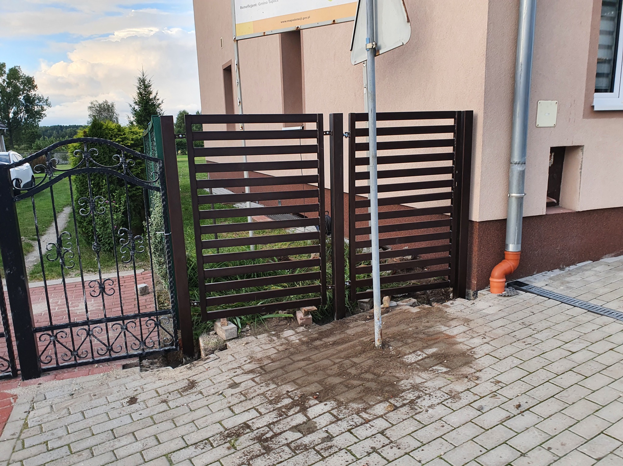 Nowoczesna, pozioma brama z brązowych profili, z widocznym zawiasem, zamontowana obok klasycznej, czarnej, metalowej bramy z ozdobnymi elementami.