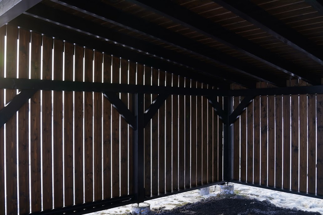 Konstrukcja z ciemnego drewna z pionowymi deskami i czarnymi belkami, widok od wewnątrz, z fragmentami śniegu na ziemi. Oświetlenie słoneczne przebija przez szczeliny.