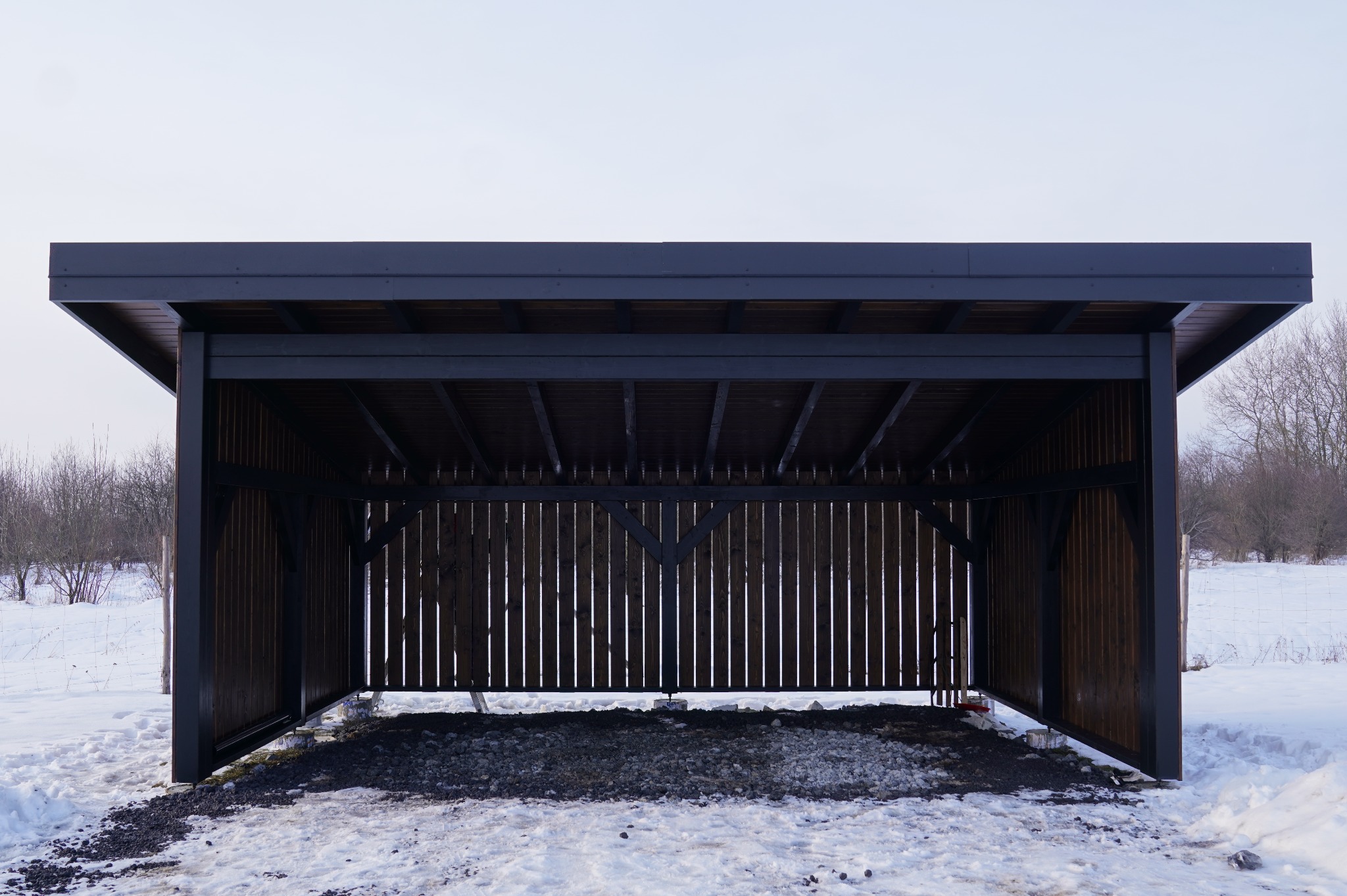 Nowoczesna, ciemna konstrukcja z drewna, prawdopodobnie wiata lub zadaszenie, na tle zimowego krajobrazu z drzewami i śniegiem. Minimalistyczny design.