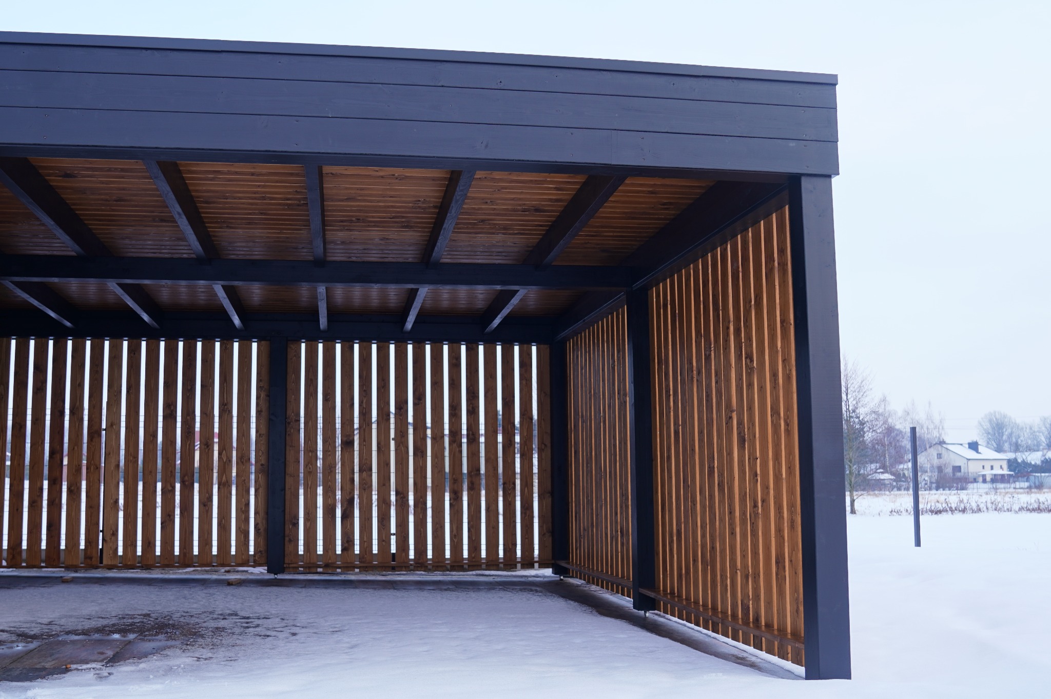 Nowoczesna, drewniana konstrukcja z ciemnymi elementami, prawdopodobnie pergola, w zimowym krajobrazie. Widoczne pionowe deski i drewniany sufit.