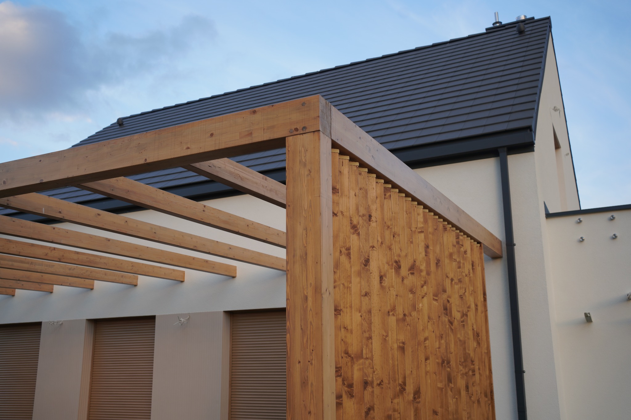 Drewniana pergola z pionowymi lamelami, przylegająca do jasnego budynku z ciemnym dachem. Widoczne elementy konstrukcyjne i detale wykończenia.