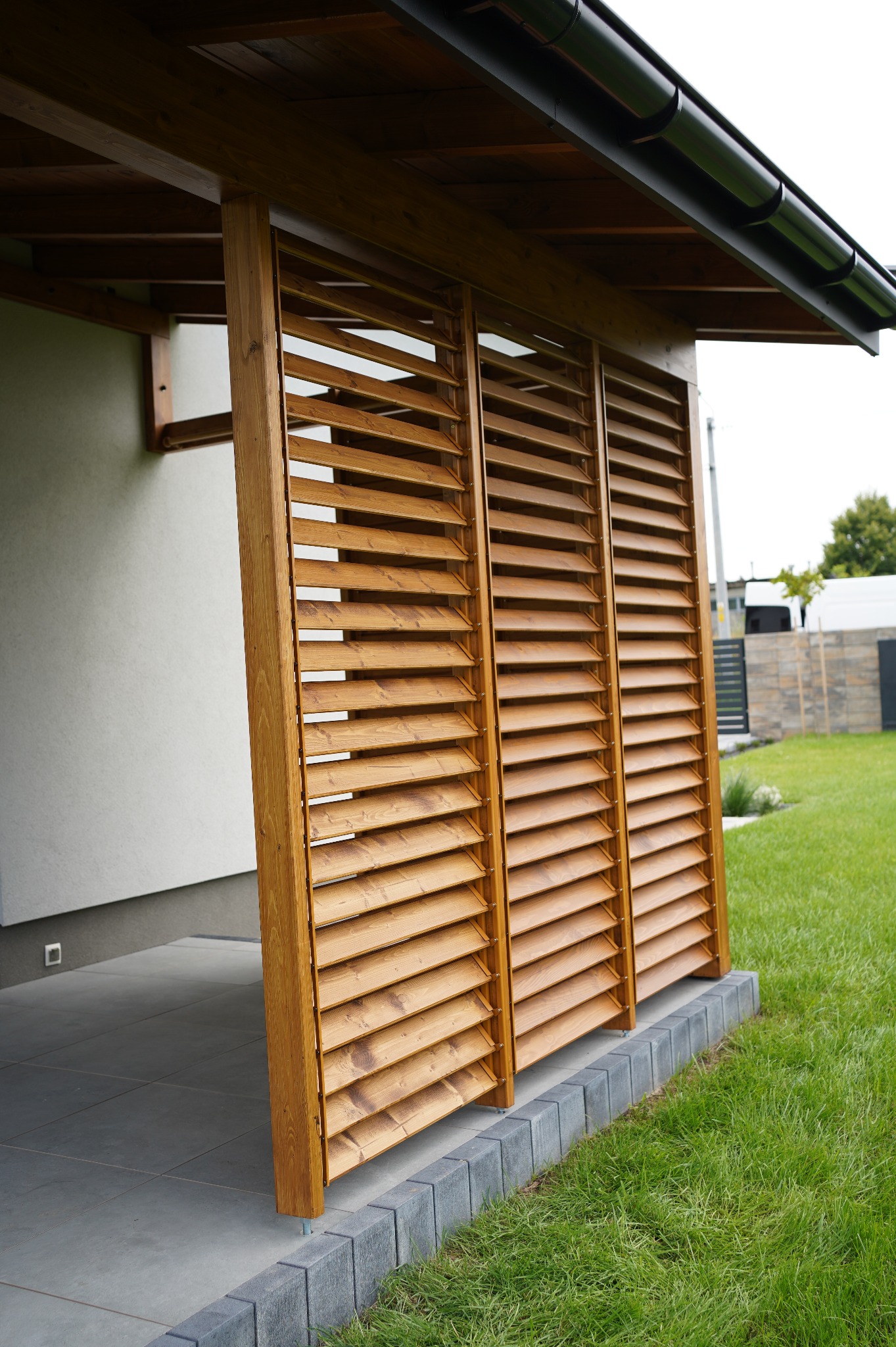 Drewniana zabudowa z ruchomych żaluzji, tworząca przesłonę na tarasie. Widoczne elementy konstrukcyjne i detale wykończenia. Ujęcie z bliska, zieleń trawnika w tle.