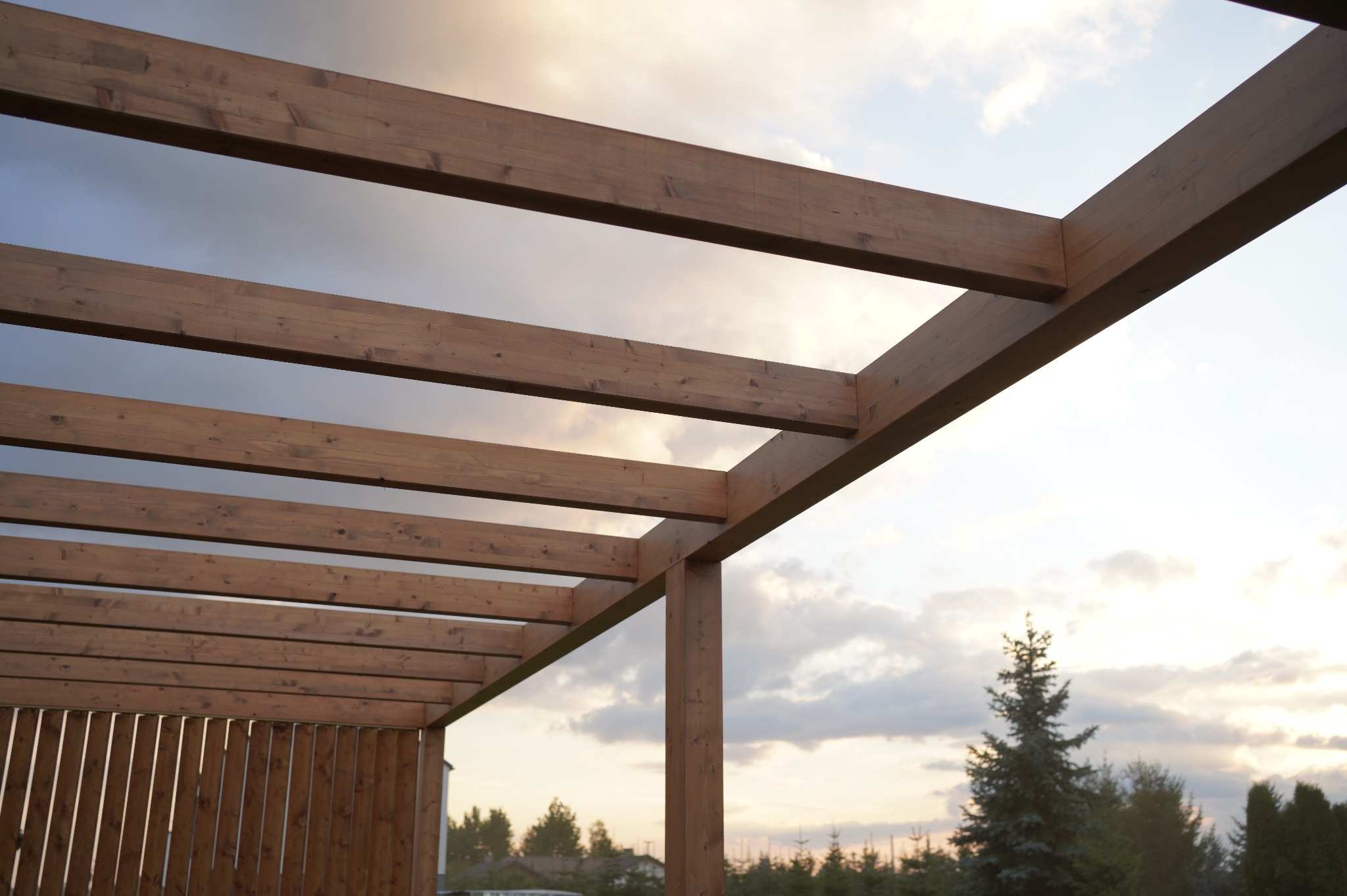 Drewniana konstrukcja pergoli z widocznymi belkami na tle nieba o zmierzchu. Elementy konstrukcyjne z bliska, ukazujące detale połączeń i fakturę drewna.