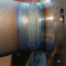 Tomasz Wcisło - Precyzyjne spawy na metalowym elemencie z widocznymi tęczowymi przebarwieniami termicznymi, ukazujące umiejętności spawacza w zbliżeniu.