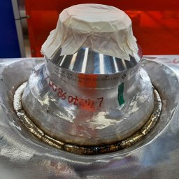 Tomasz Wcisło - Precyzyjne spawanie kołnierza rury ze stali nierdzewnej, zabezpieczonego papierem, z widocznymi śladami obróbki i oznaczeniami technicznymi. Detal spawalniczy.