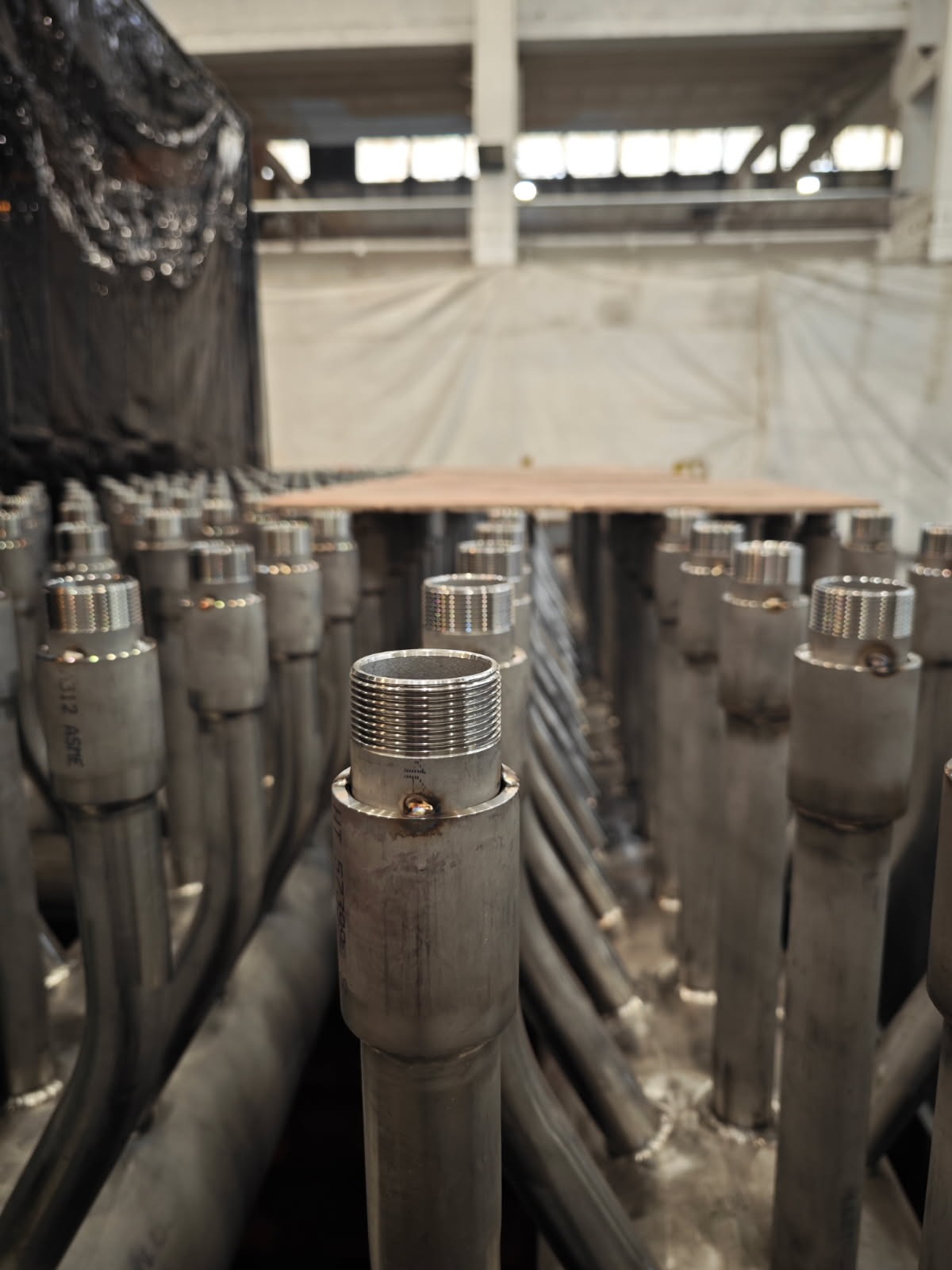 Rząd metalowych rur z gwintowanymi końcówkami, spawanych ze sobą, w hali produkcyjnej. Widoczne ślady spawania i oznaczenia na rurach.