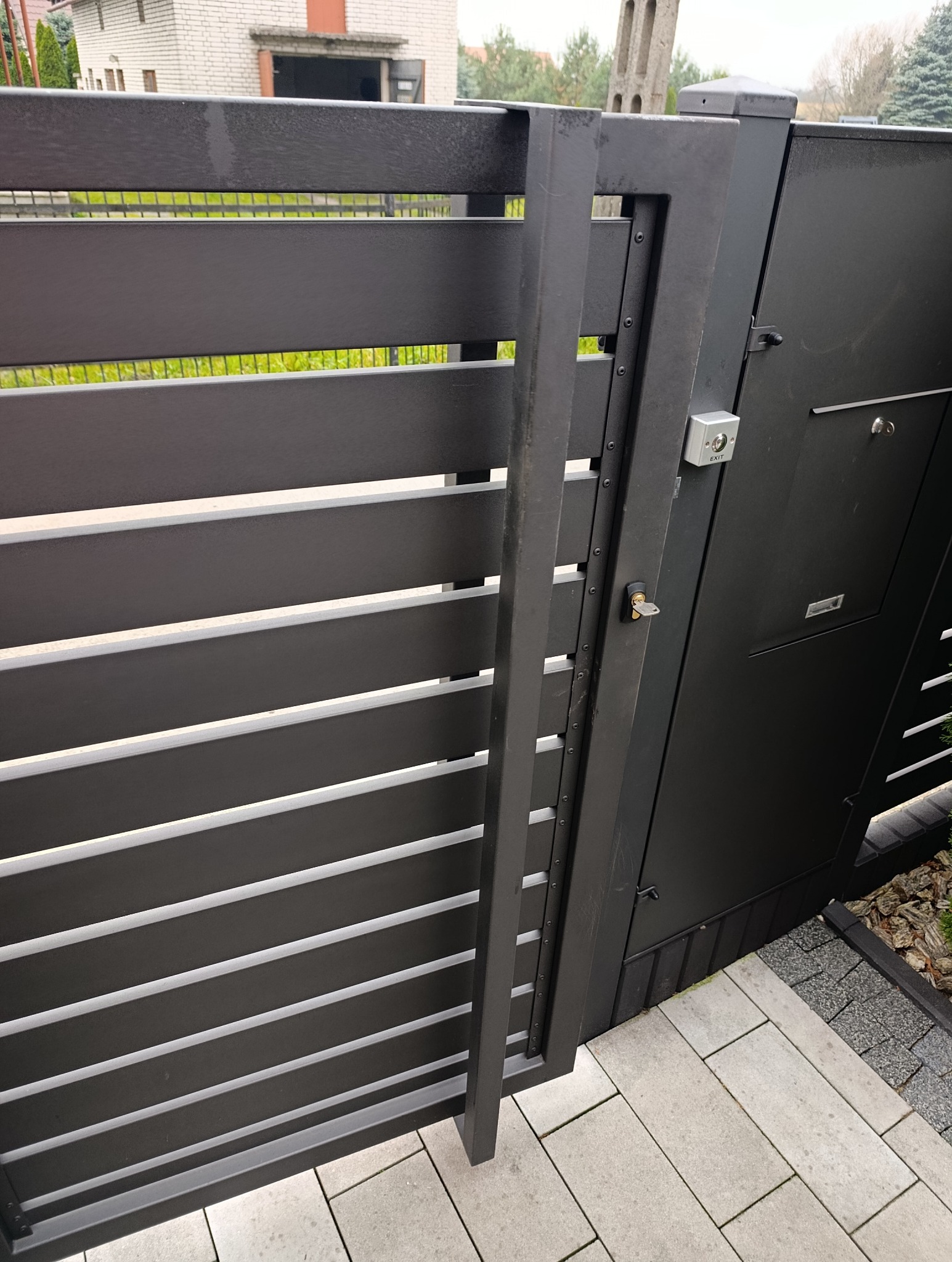 Nowoczesna, pozioma brama wjazdowa z czarnego metalu, z widocznym domofonem i skrzynką na listy, osadzona na szarej kostce brukowej.