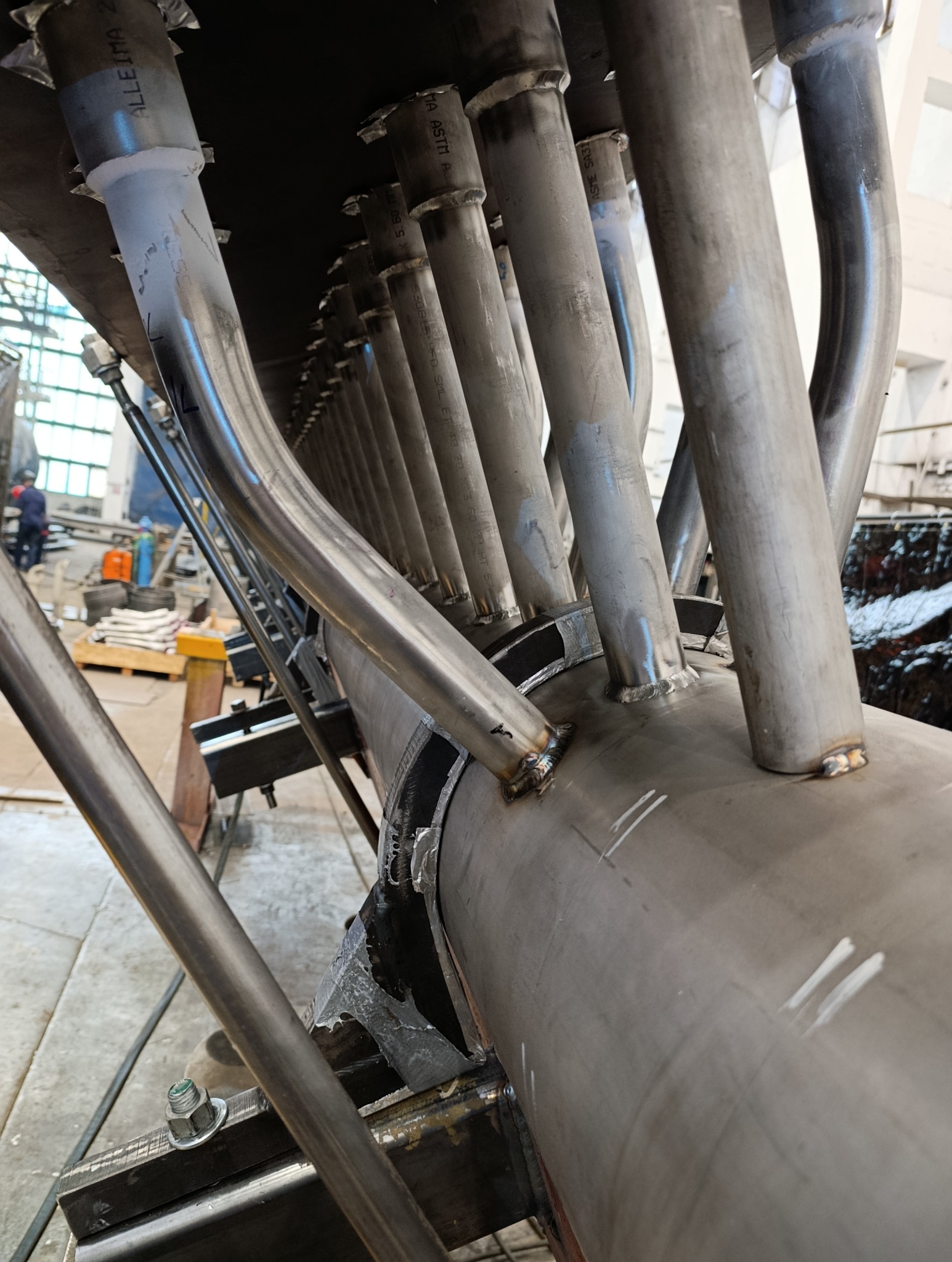 Detal spawanej konstrukcji stalowej z rur, widoczne spawy i oznaczenia na metalu. W tle warsztat z elementami wyposażenia i pracownik w oddali.