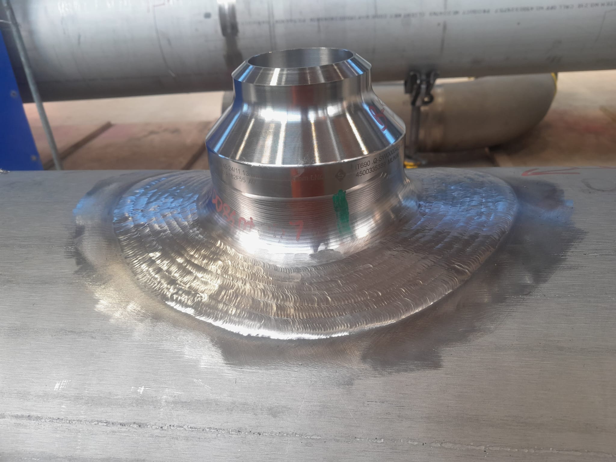 Polerowane, metalowe złącze rurowe, spawane do rury. Widoczne ślady po spawaniu i polerowaniu. Złącze z oznaczeniami producenta.