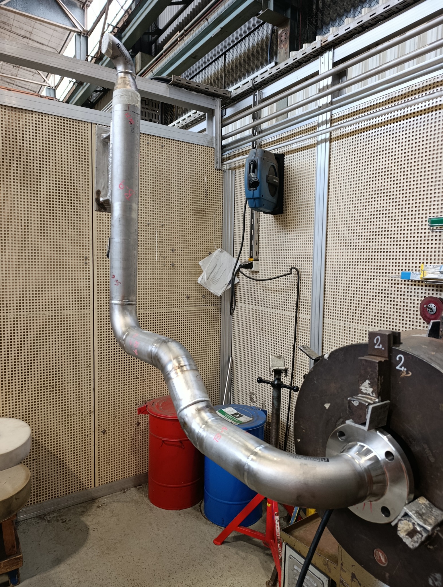 Metalowa rura z flanszą, zamocowana w uchwycie tokarskim, widoczne ślady spawania i oznaczenia. W tle warsztat z perforowanymi ścianami i narzędziami.