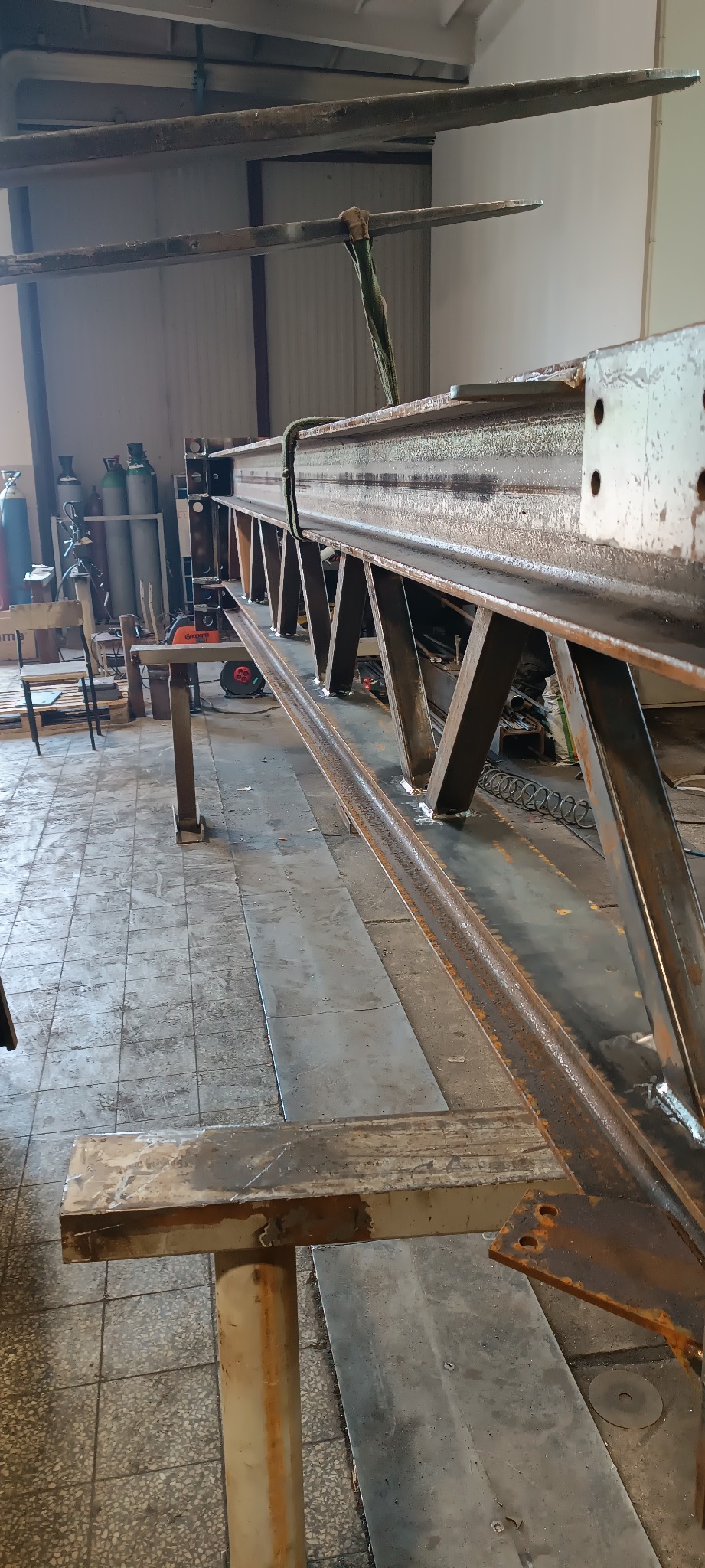 Surowa, spawana konstrukcja stalowa w warsztacie, z widocznymi spawami i elementami mocującymi. W tle butle z gazami technicznymi i narzędzia.