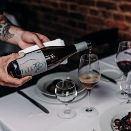 Wine Way Ticket - Nalewanie białego wina z butelki Papillon Macération Sauvignon 2021 do kieliszka na stole z białym obrusem, w tle ceglana ściana i dania.