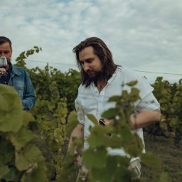 Wine Way Ticket - Degustacja w winnicy: Mężczyzna w białej koszuli ocenia wino, drugi degustuje z kieliszka. Krzewy winorośli w tle, pochmurne niebo.