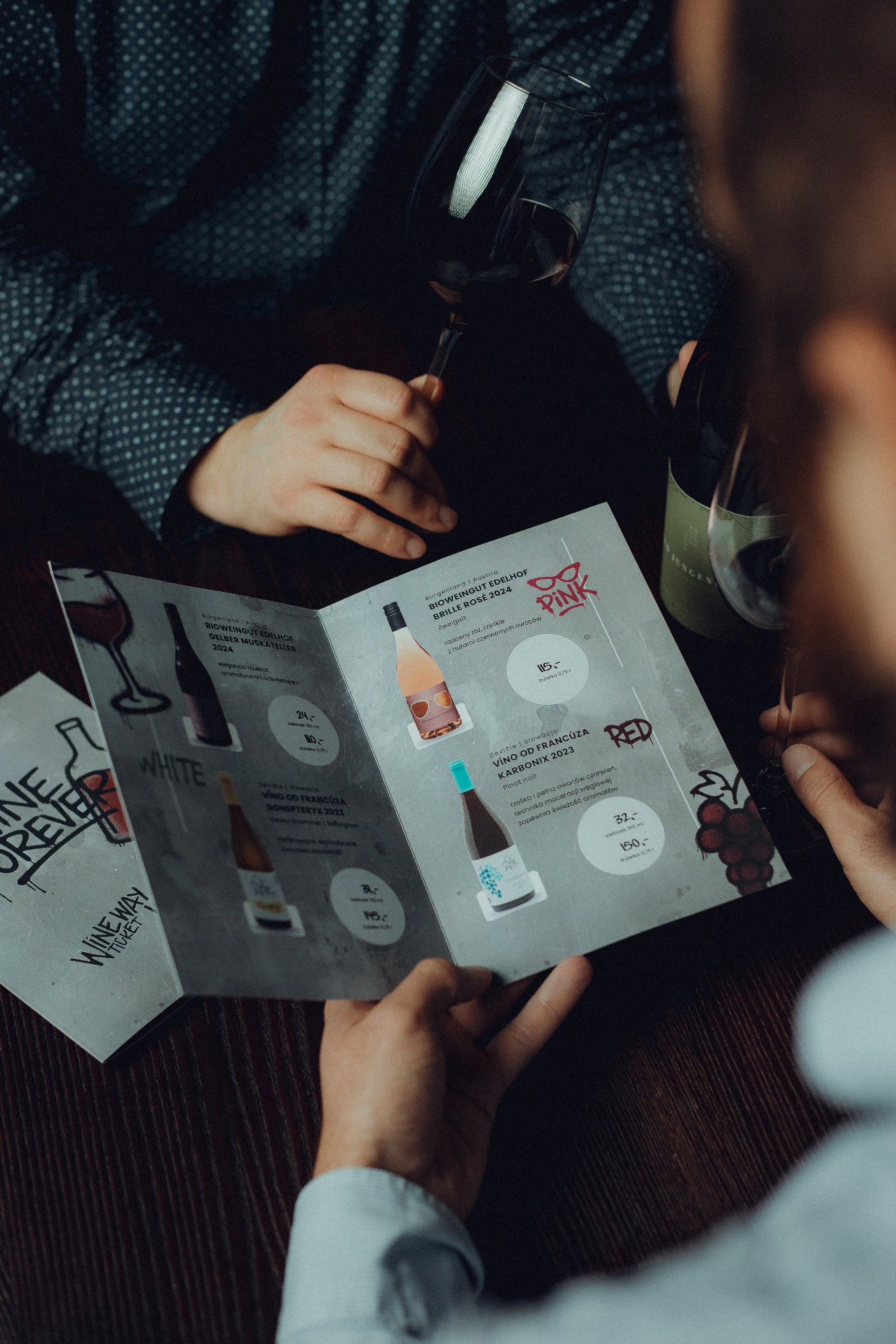 Osoby przeglądają kartę win z ofertą win białych, różowych i czerwonych, w tle kieliszki i butelka wina. Degustacja wina w lokalu.