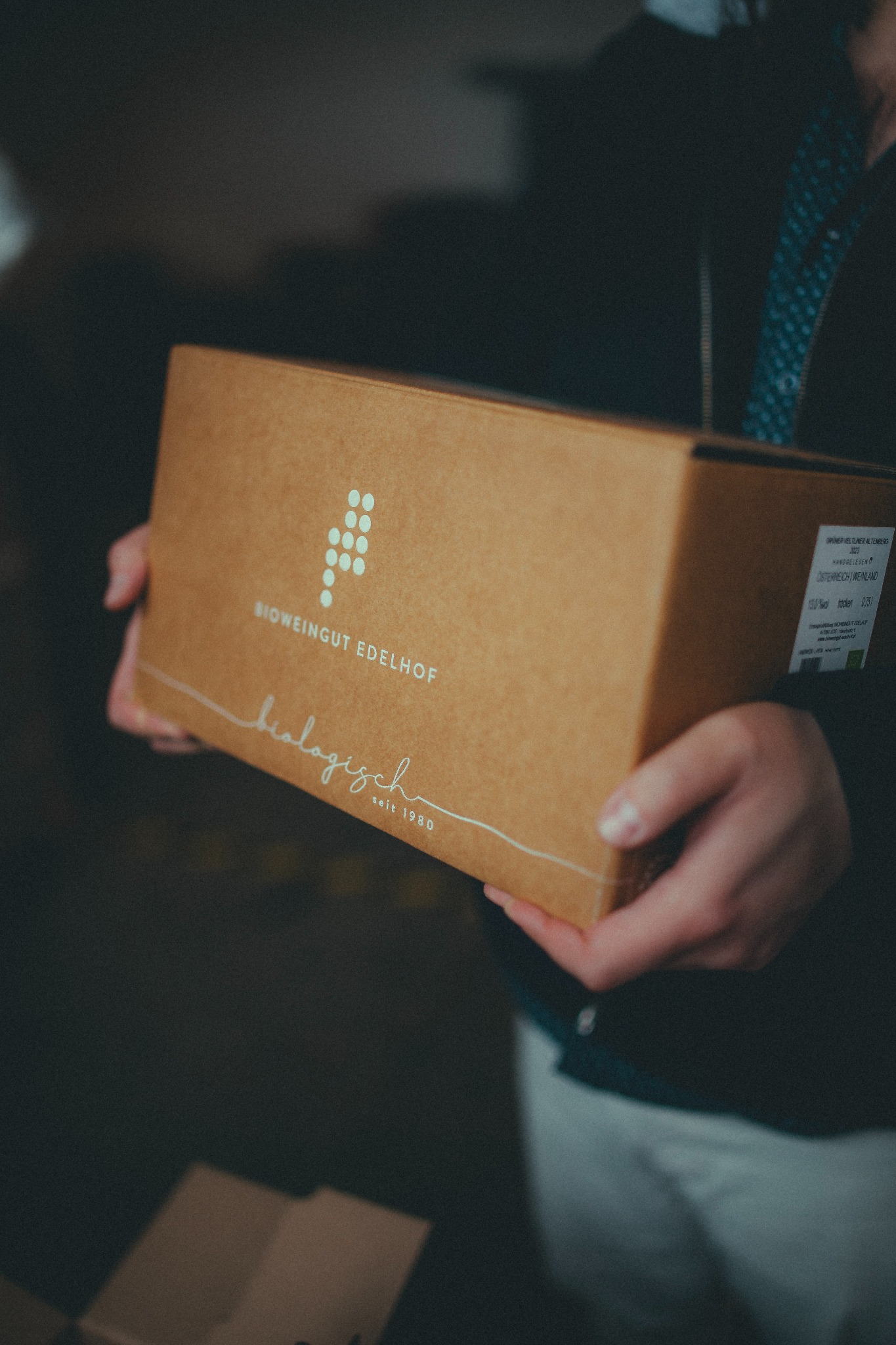 Osoba trzyma kartonowe pudełko z logo Bioweingut Edelhof, prezentujące ekologiczne wino z Austrii, rocznik 2023, z widoczną etykietą informacyjną.