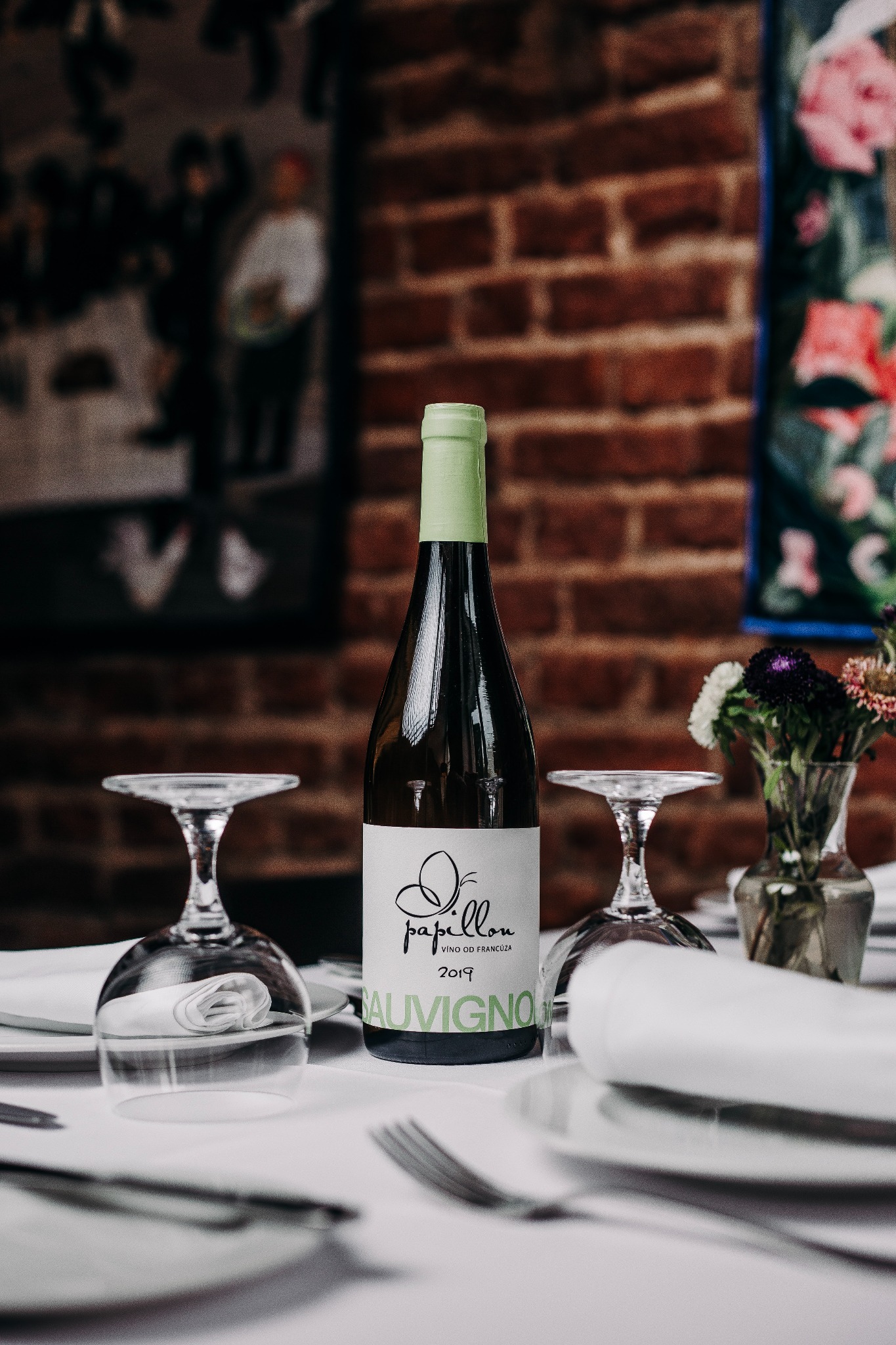 Butelka białego wina Sauvignon Blanc 2019 na stole z nakryciem, kieliszki do góry dnem, w tle ceglana ściana i kwiaty w wazonie. Elegancka aranżacja.