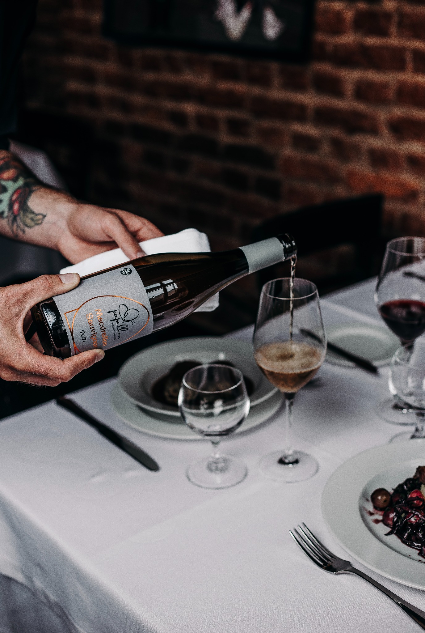 Nalewanie białego wina z butelki Papillon Macération Sauvignon 2021 do kieliszka na stole z białym obrusem, w tle ceglana ściana i dania.